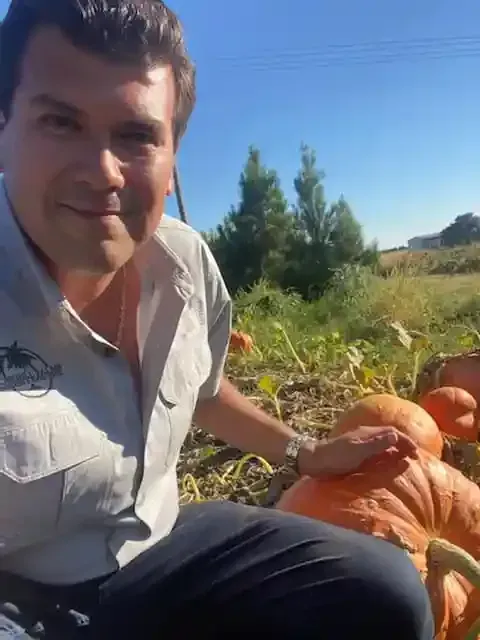 Man kneeling beside a large pumpkin in a sunny field, smiling at the camera