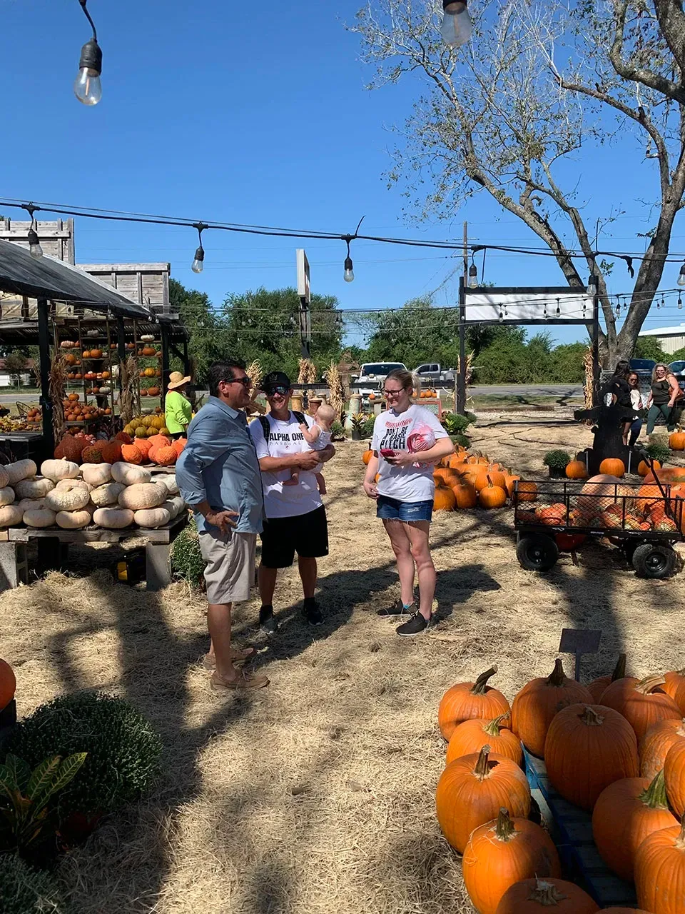 Three people stand among pumpkins at an outdoor fall market under a clear blue sky.
