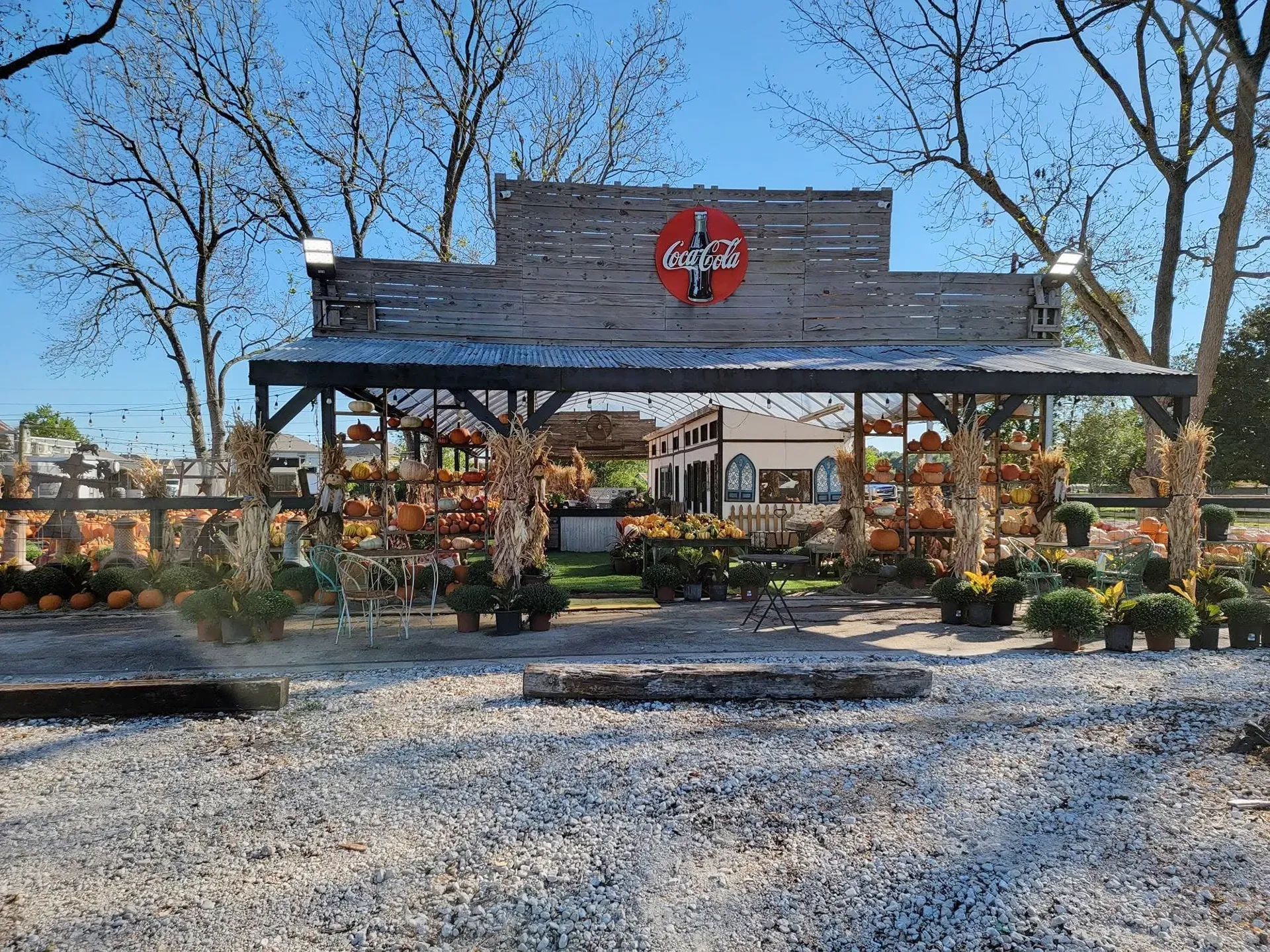 Rustic outdoor market stand with a red sign, potted plants, and gravel foreground under a clear blue sky