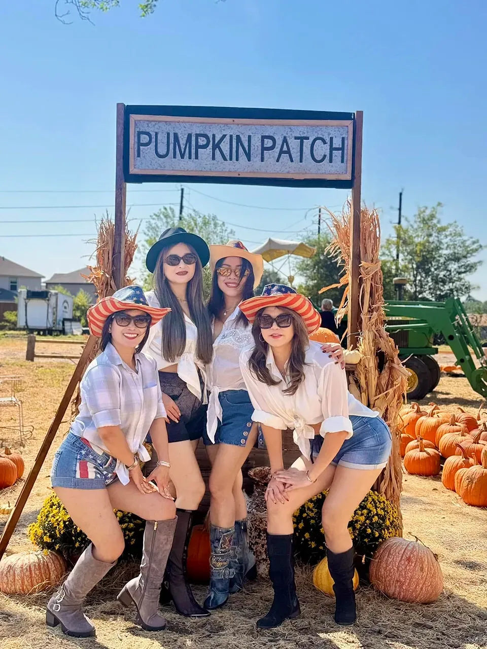 Four women pose under a Pumpkin Patch sign, surrounded by pumpkins and a tractor on a sunny farm.