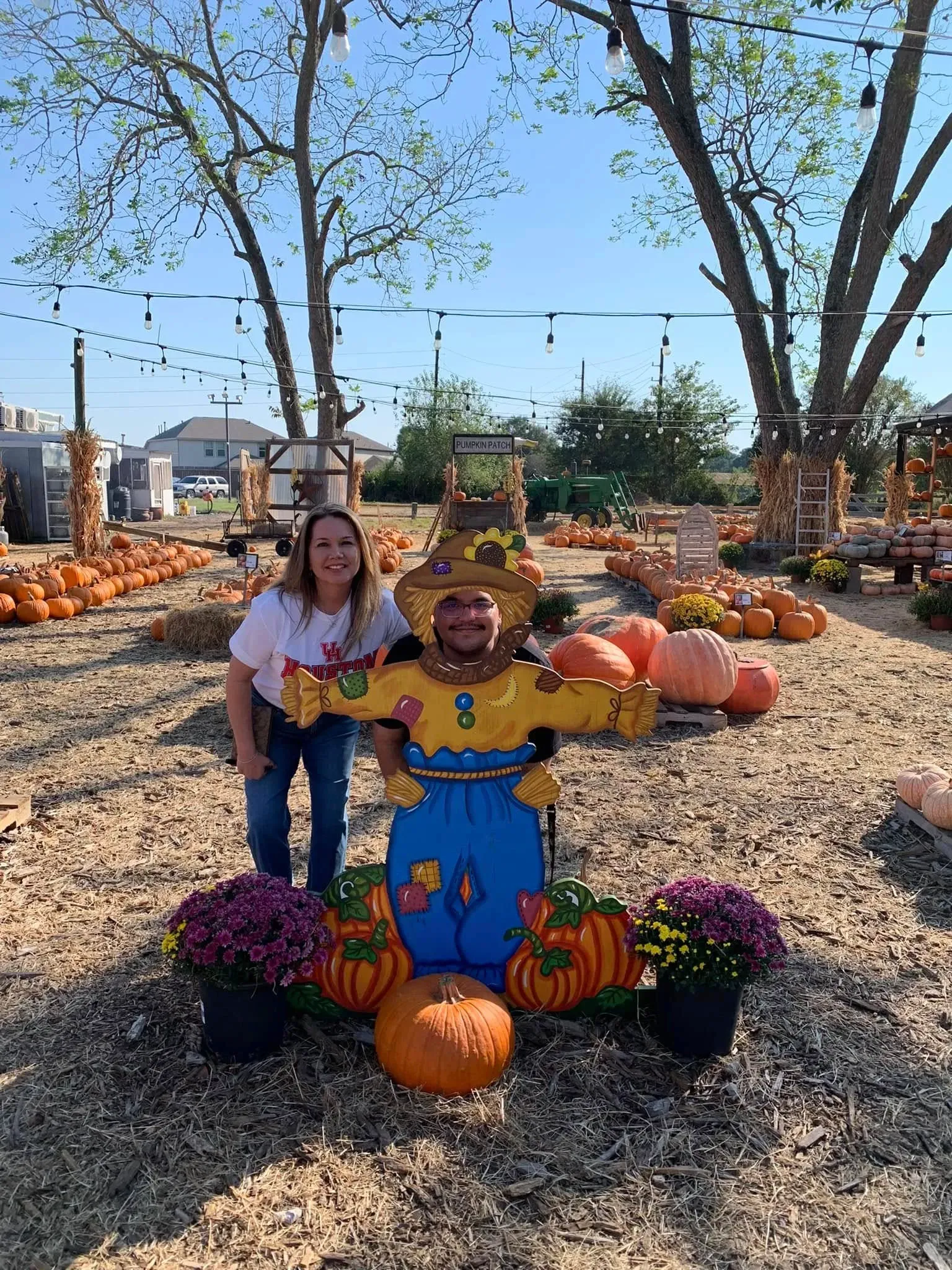 Woman posing beside a colorful scarecrow and pumpkins at a fall festival outdoors.