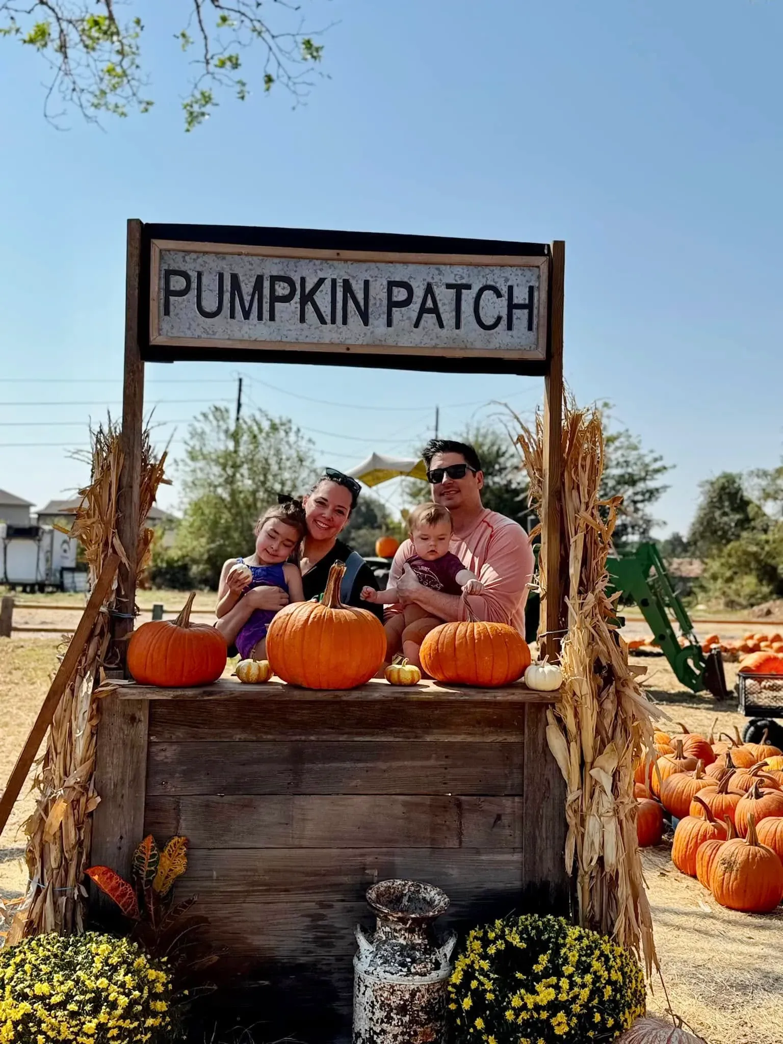 Family sitting on a wooden pumpkin patch cart with orange pumpkins and flowers under a “PUMPKIN PATCH” sign