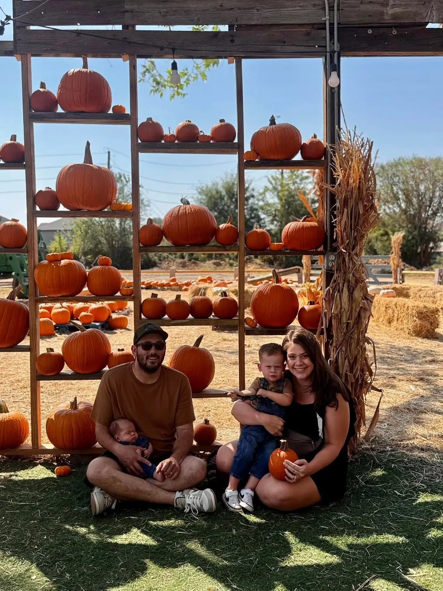 Two people are sitting on grass in front of a pumpkin display at an outdoor fall market.