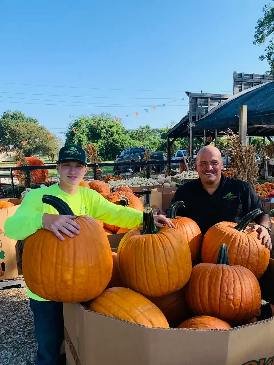 Two people posing with large pumpkins at an outdoor autumn market under a bright blue sky