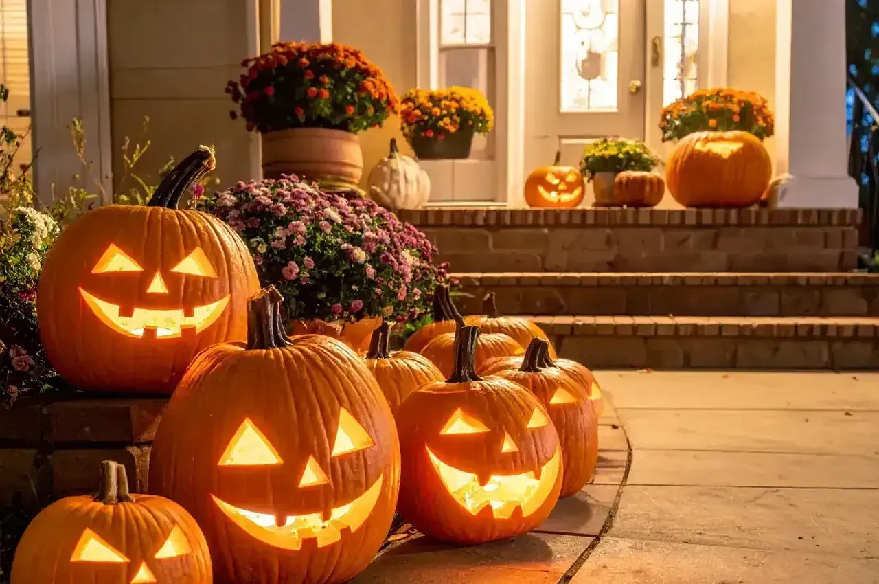 Halloween pumpkins with carved glowing faces on a porch and steps, surrounded by potted flowers at dusk