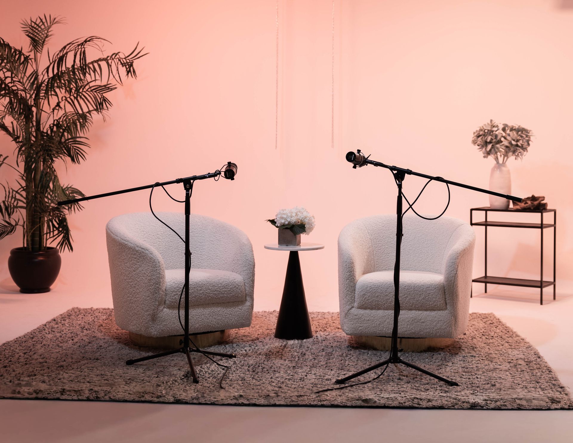 Two white armchairs with microphones on stands in a pink studio set with plants and a rug