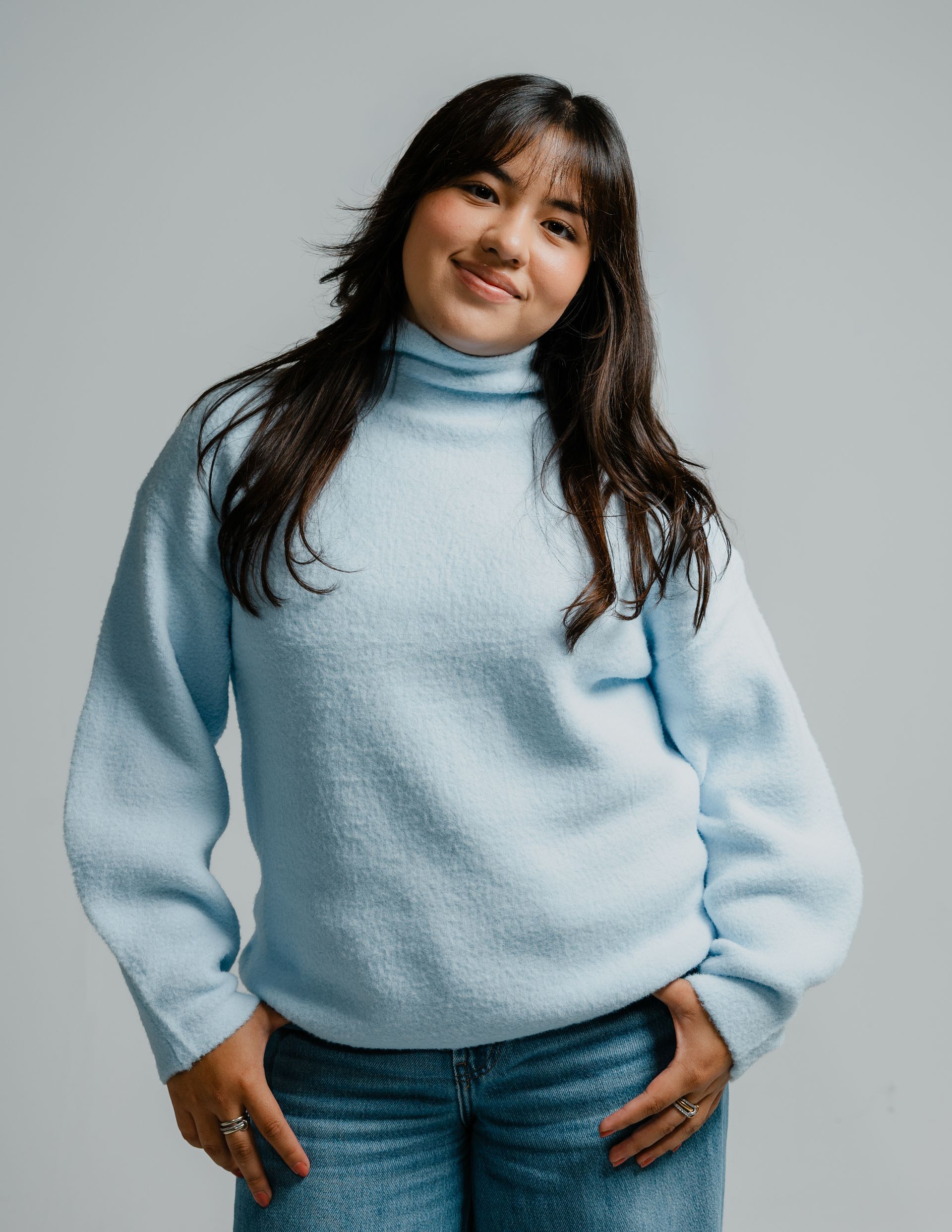 Person in a light blue sweater and jeans smiling against a gray background.