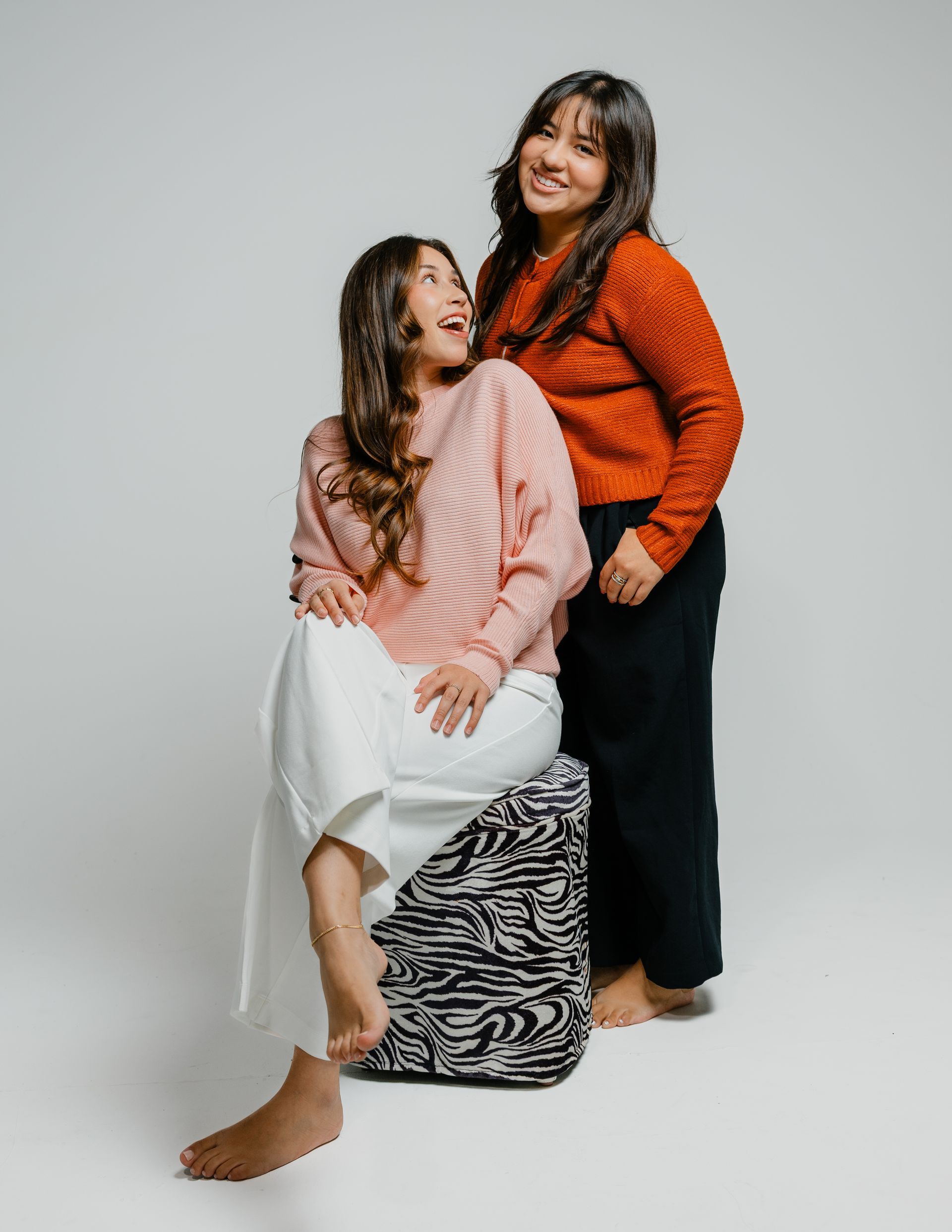 Two women pose in a studio, one seated on a patterned stool, both smiling in orange and pink tops.