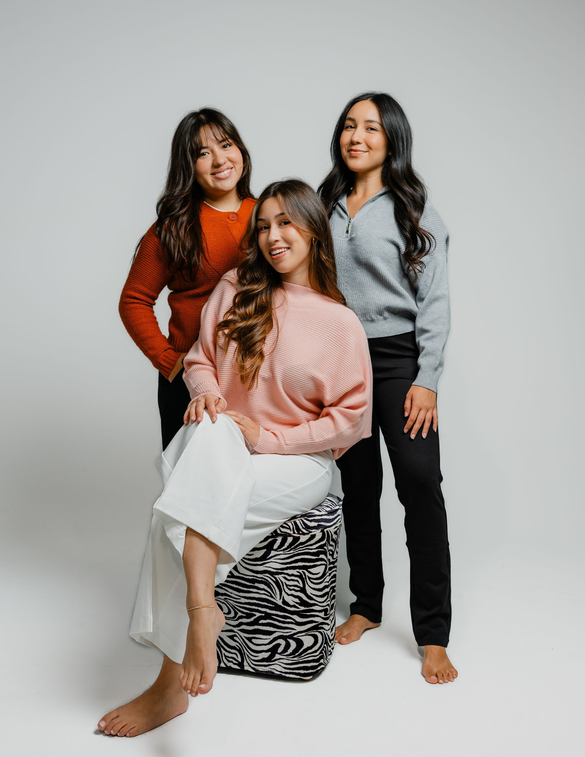 Three people posing in a studio, seated and standing in colorful sweaters and black pants.