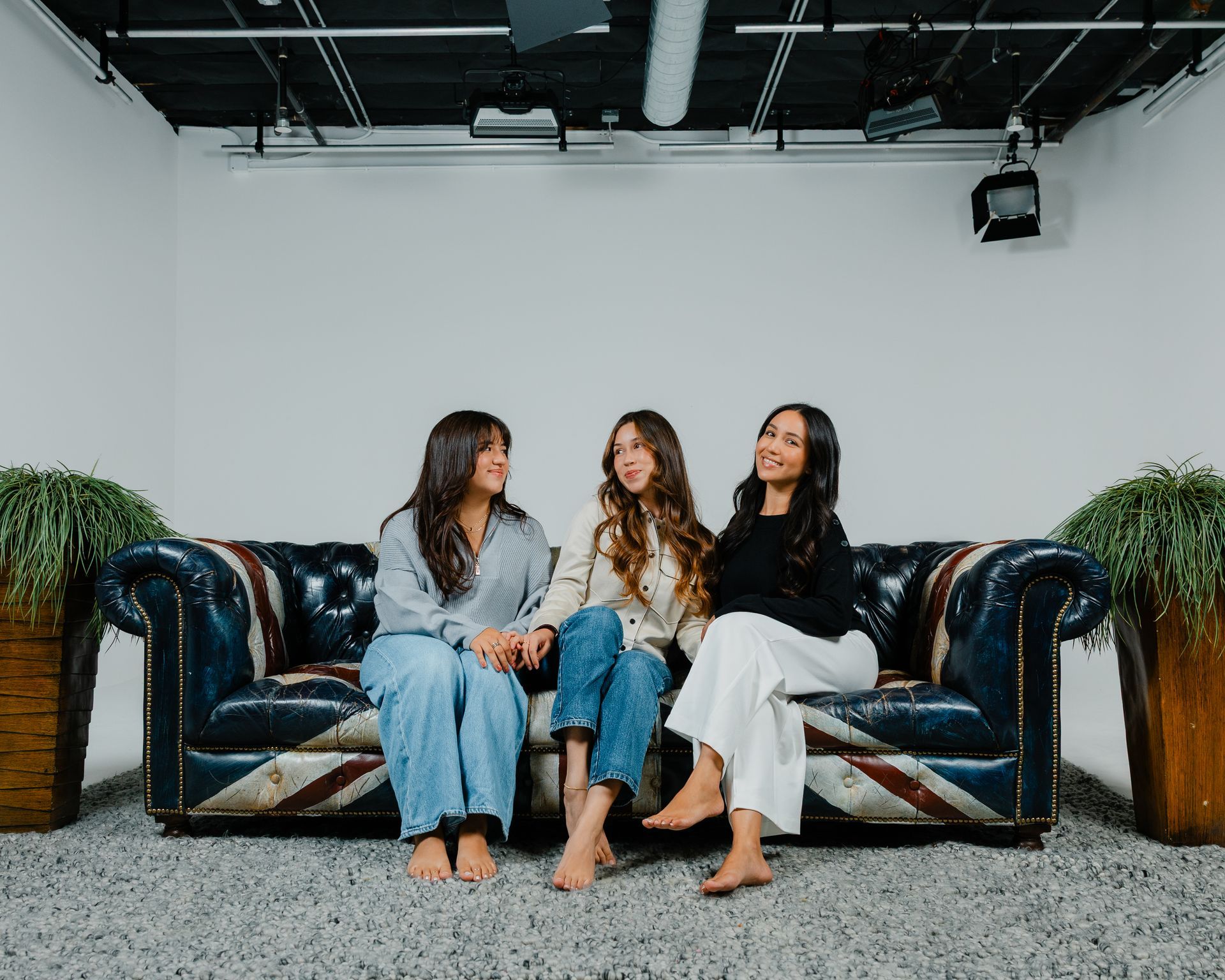 Three women sit on a blue couch in a bright studio, chatting barefoot in casual clothes.