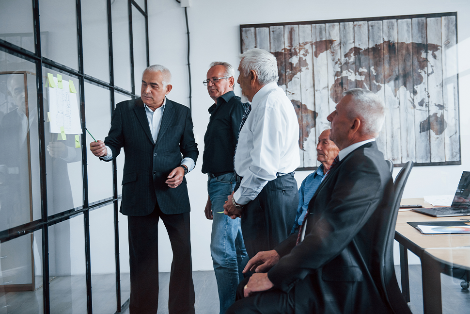 Business meeting in a glass-walled office, with one man presenting to three seated colleagues.