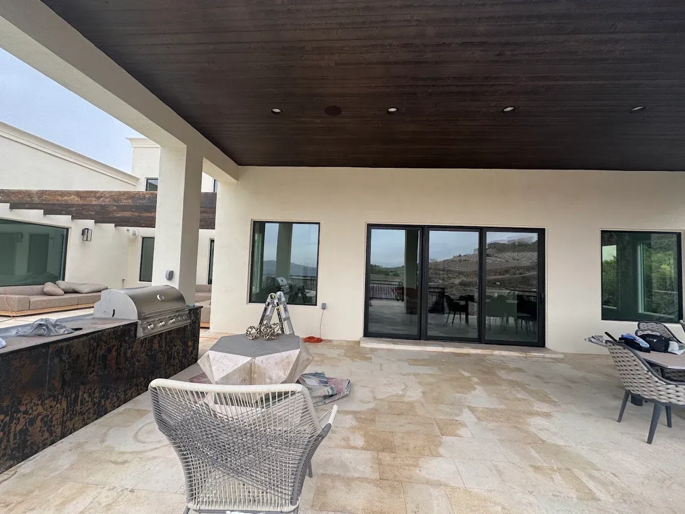Covered patio with stone floor, wicker chairs, sliding glass doors, and dark wood ceiling overlooking water.