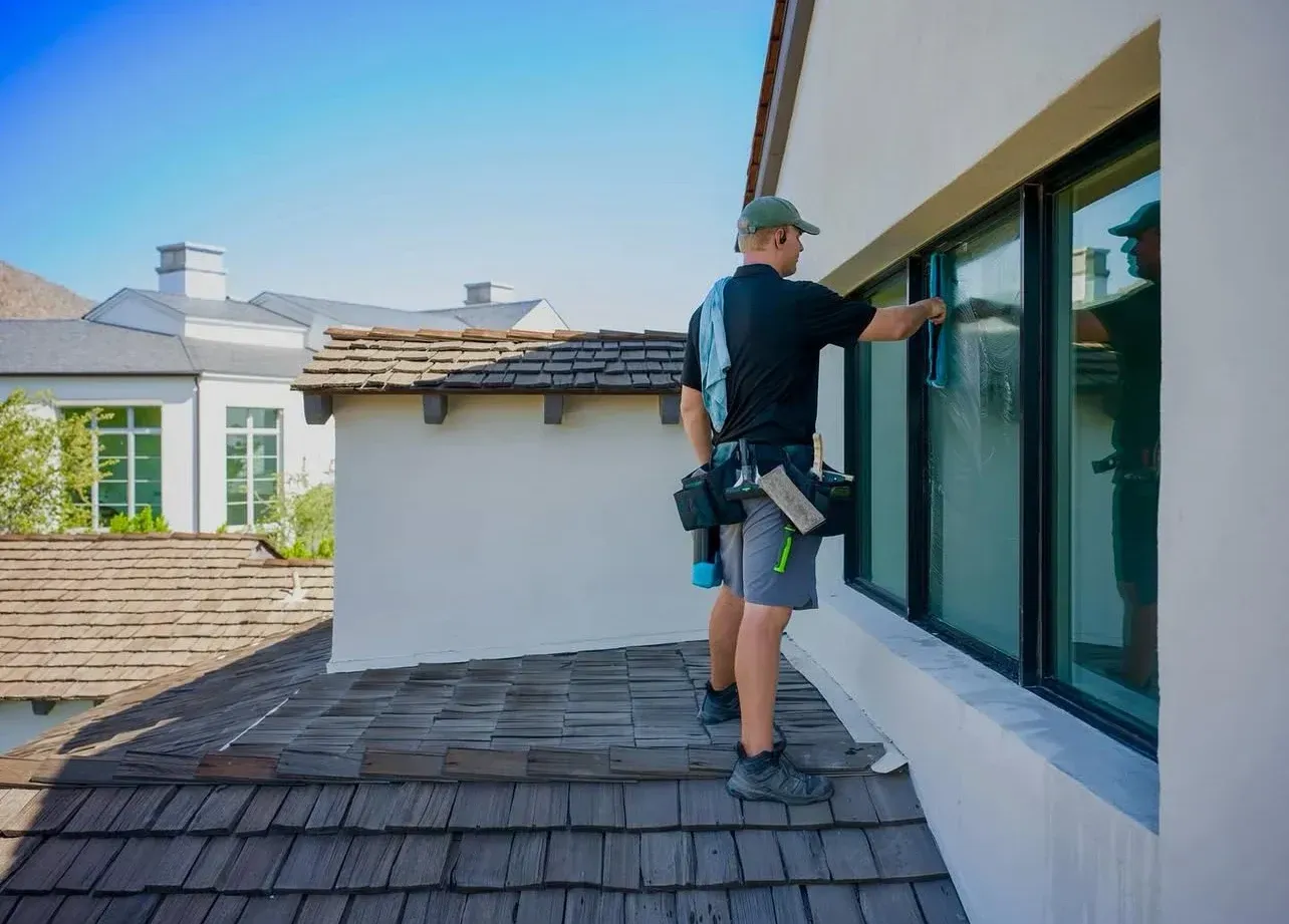 Worker cleaning a rooftop window on a house exterior under a clear blue sky