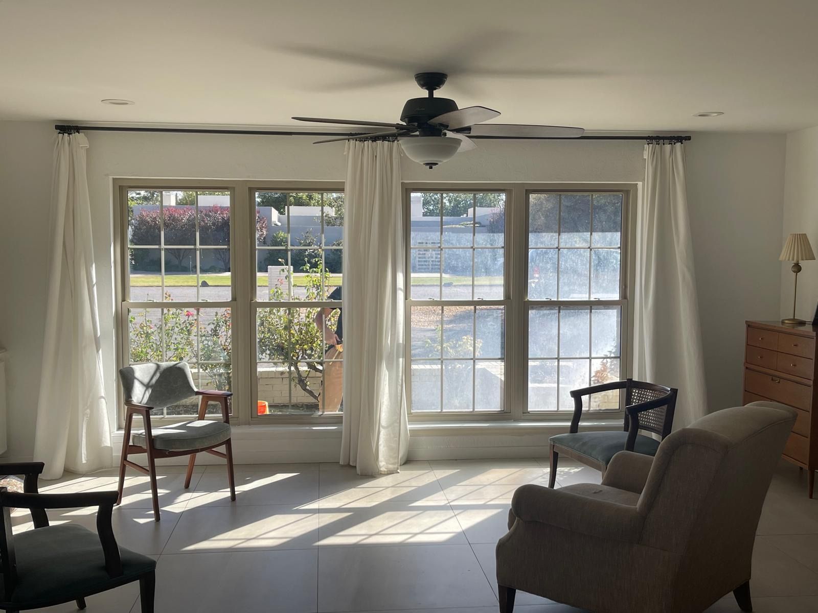 Bright living room with large windows, sheer curtains, sunlight, and chairs around a small table.
