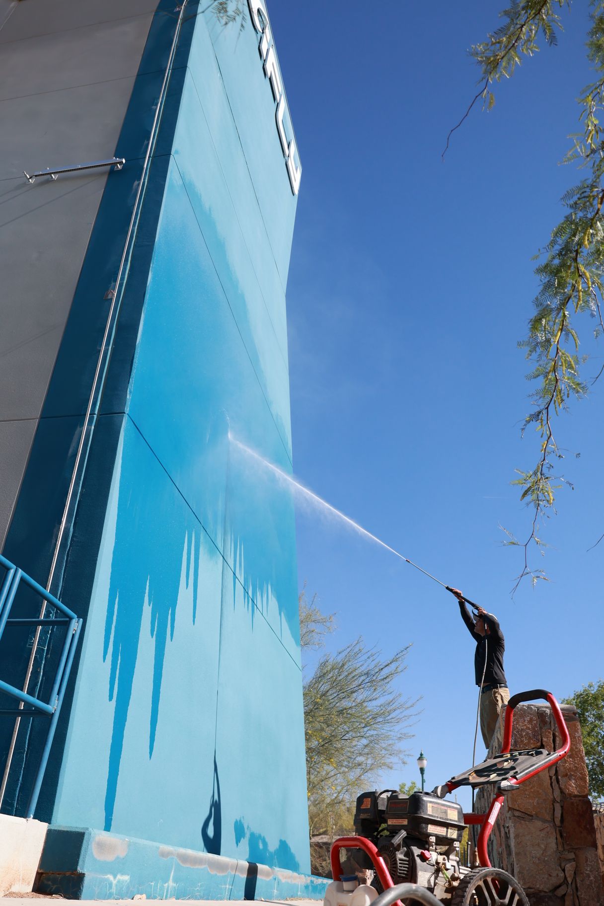 Worker power-washing a tall blue wall outdoors against a clear sky