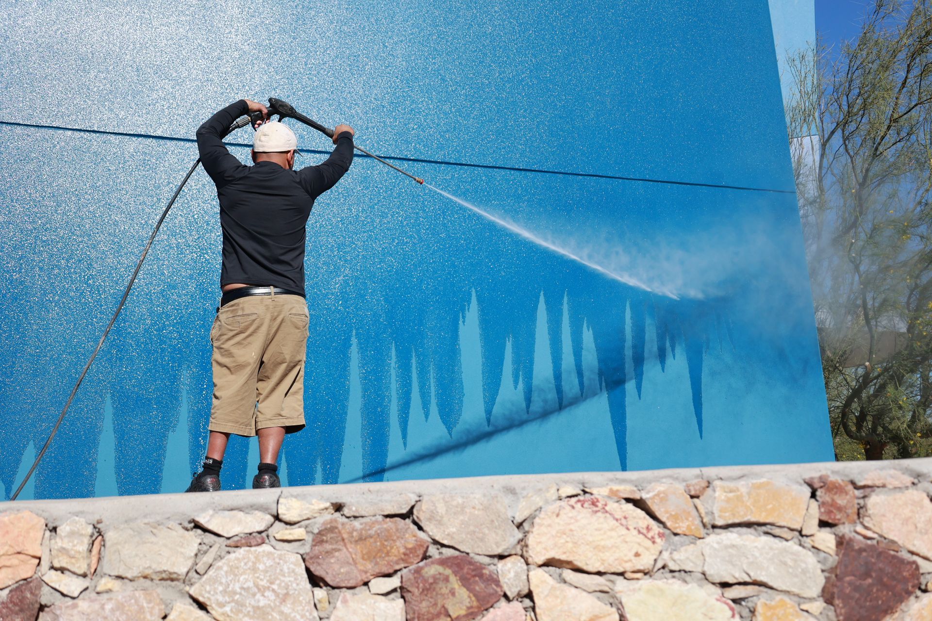 Person spraying water on a large blue mural outdoors behind a stone wall.