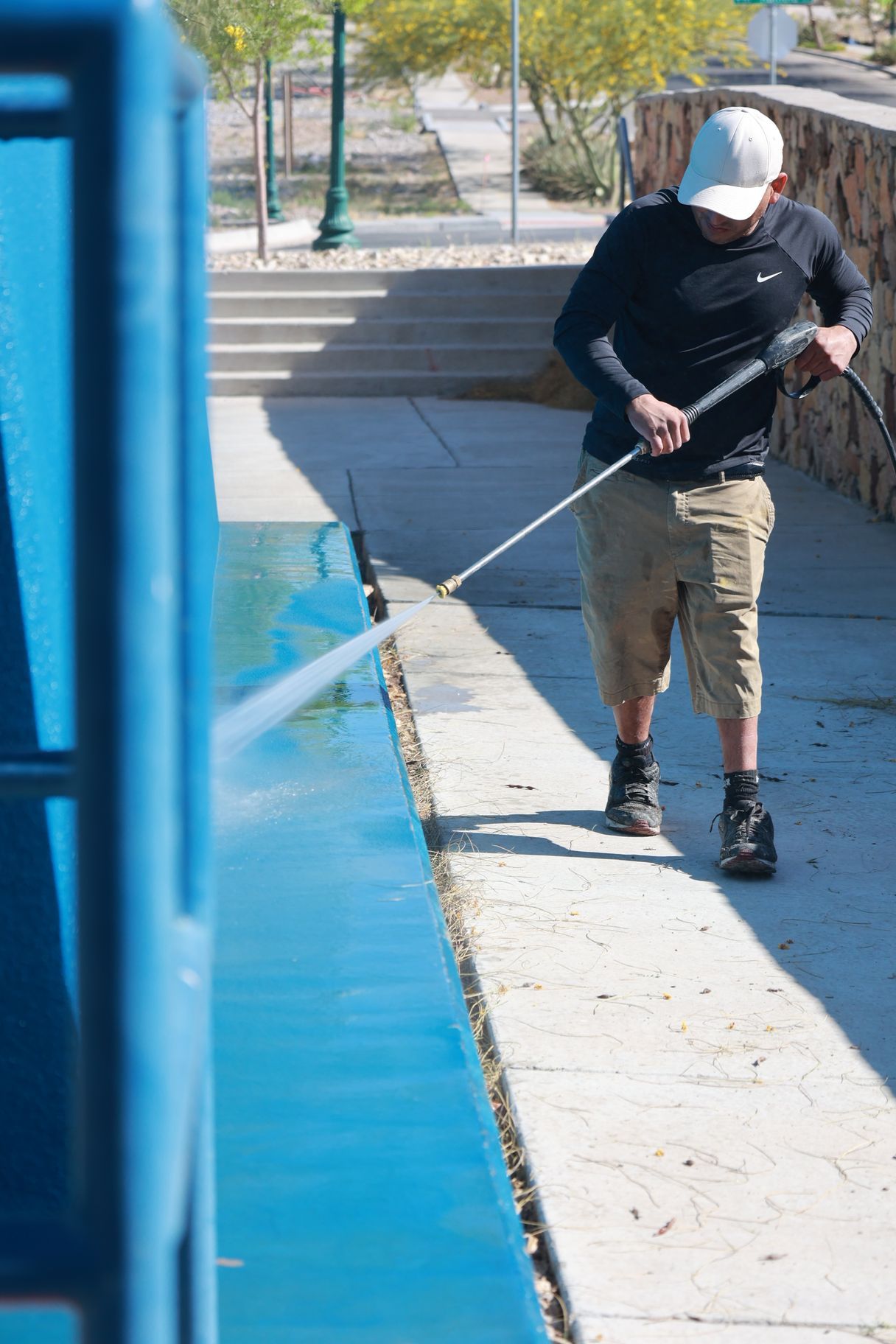 Person pressure-washing a blue wall along a sidewalk outdoors.
