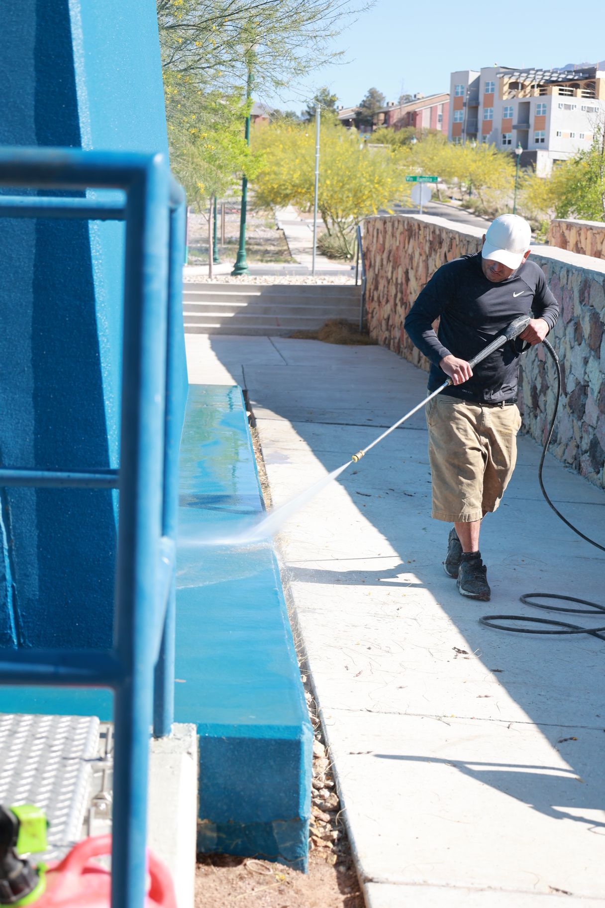 Worker pressure-washing a blue building exterior along a sunny sidewalk