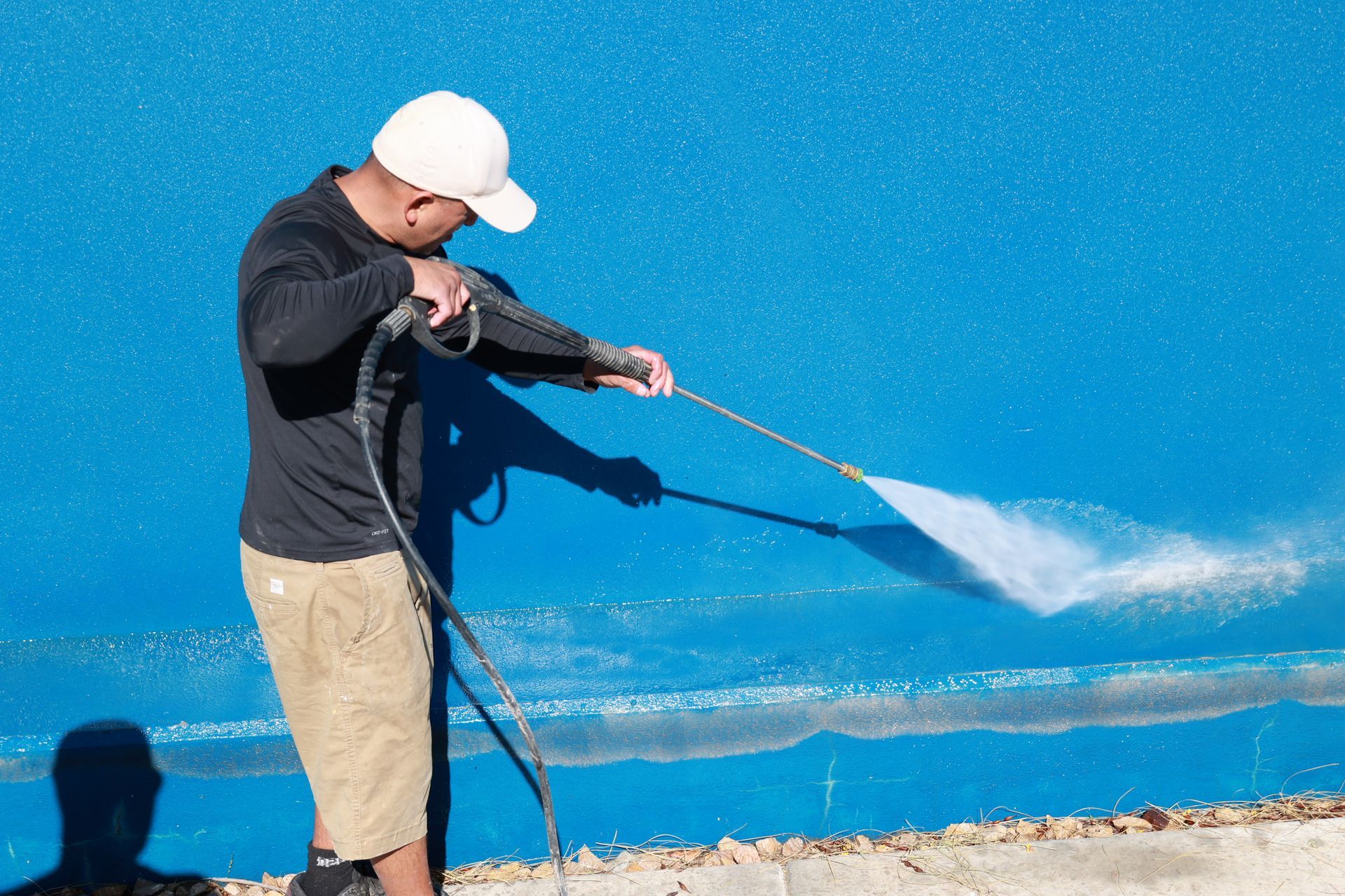 Person in a white cap pressure-washes a bright blue wall outdoors, spraying water across the surface.