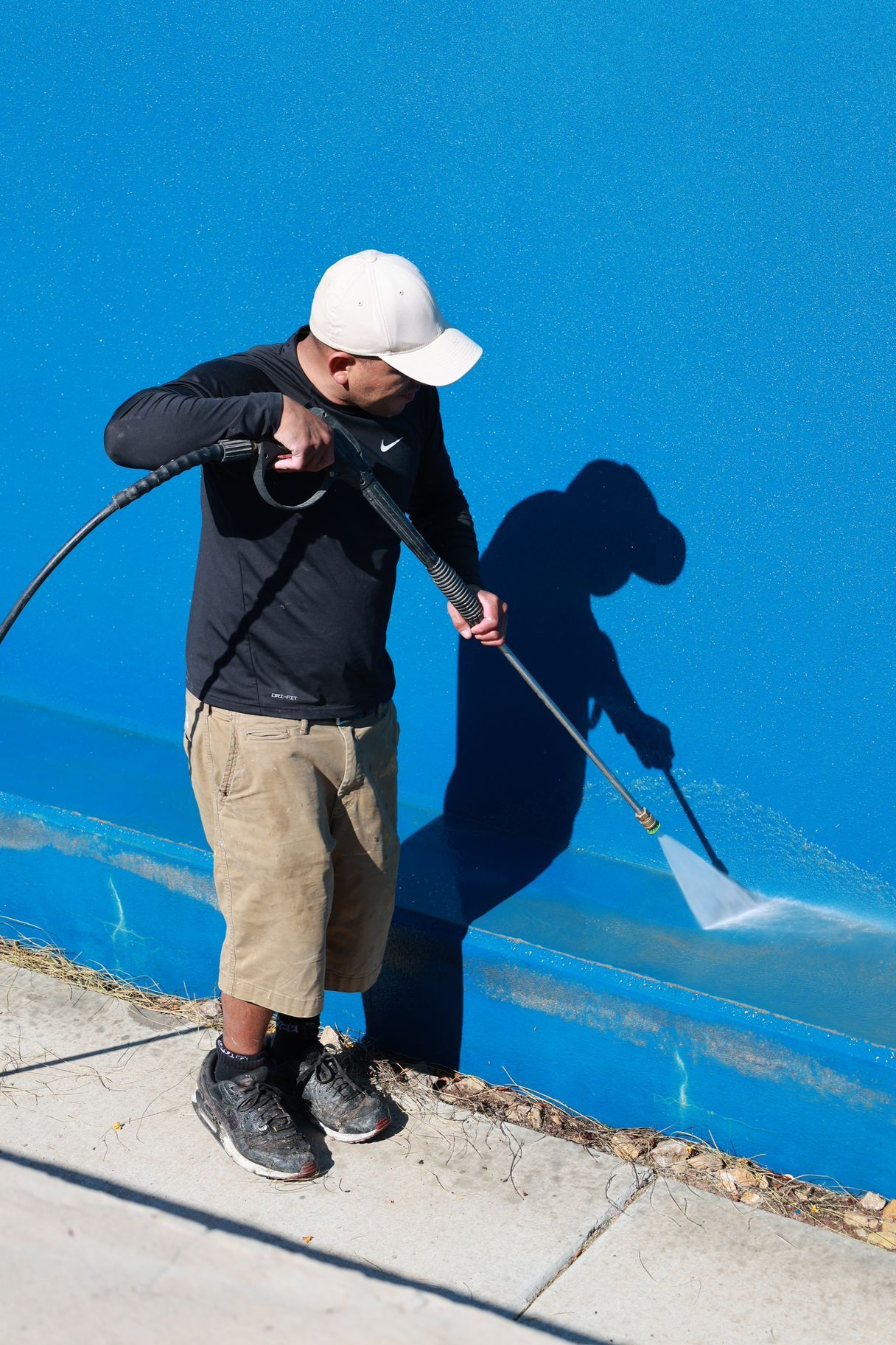 Person in white cap painting a bright blue wall with a roller, standing on a sidewalk.