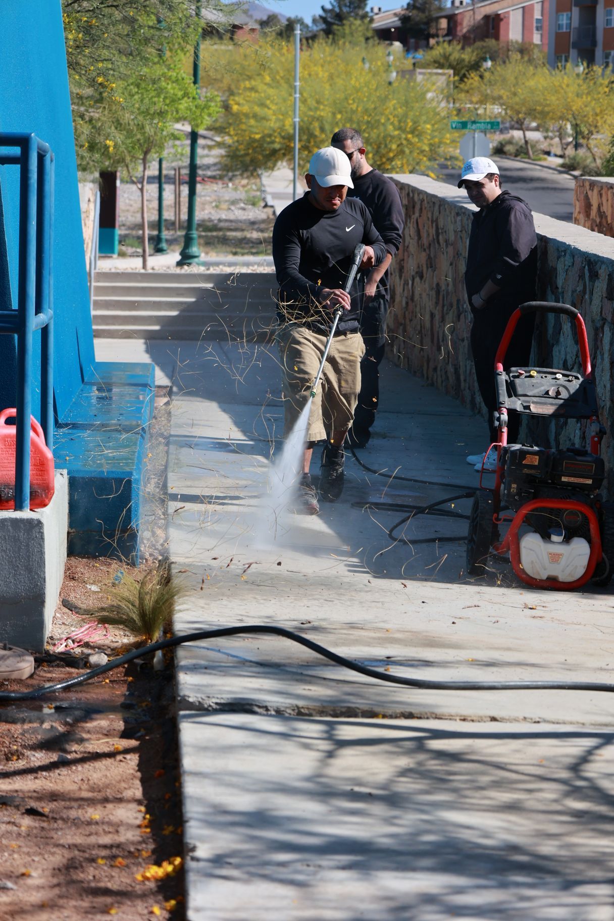 Workers using a pressure washer on a sidewalk beside a brick wall and blue building