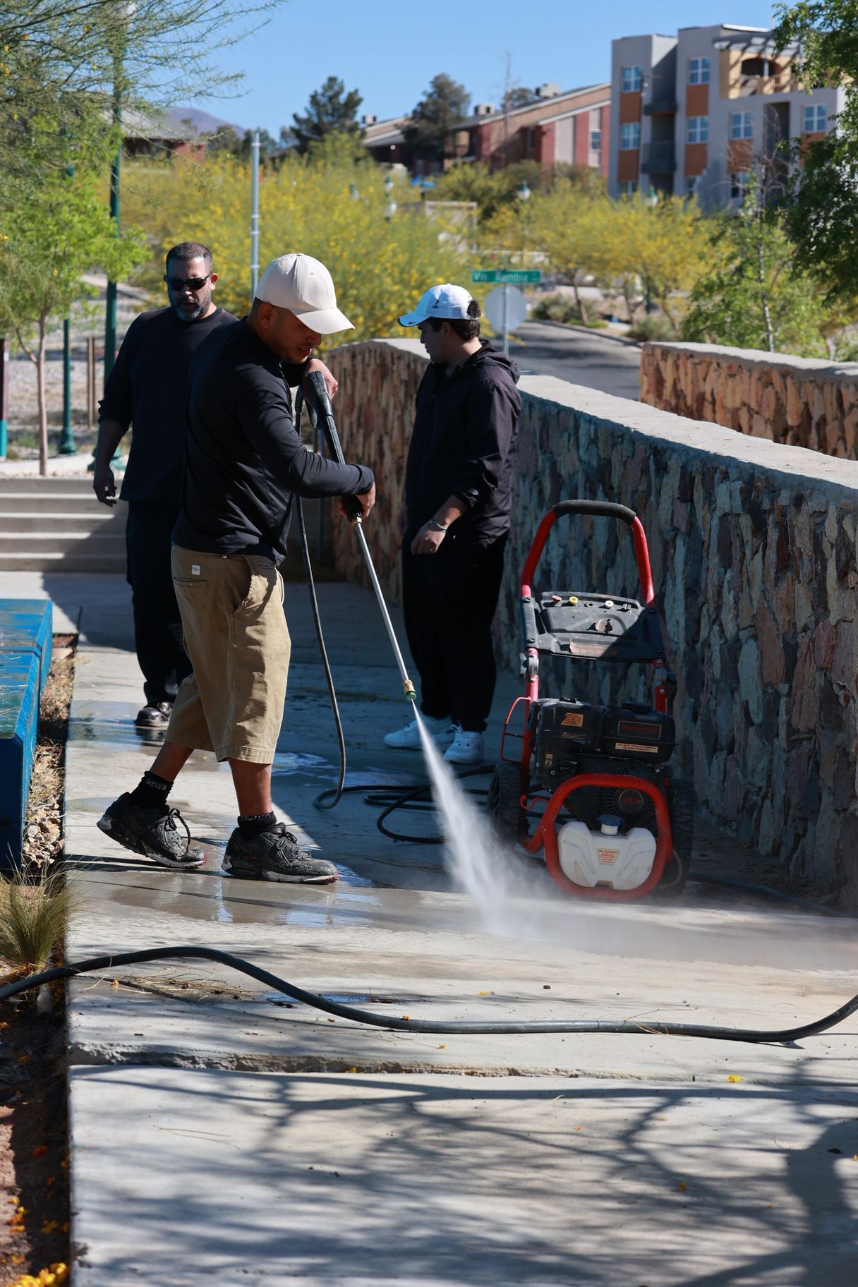 Two people pressure-wash a sidewalk beside a stone wall, with a red cart and hose nearby.