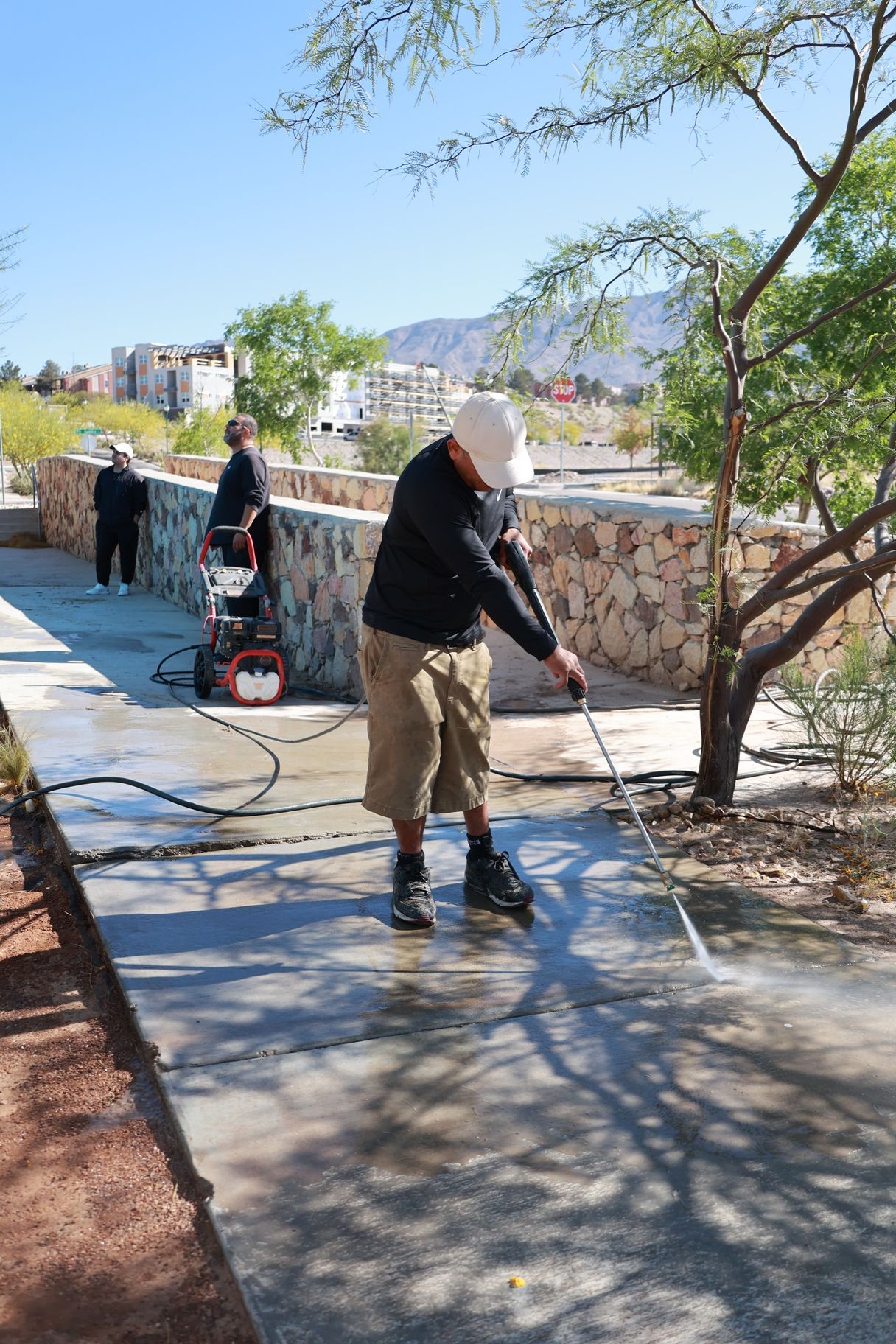 Worker pressure-washing a sidewalk beside a stone wall and trees on a sunny day