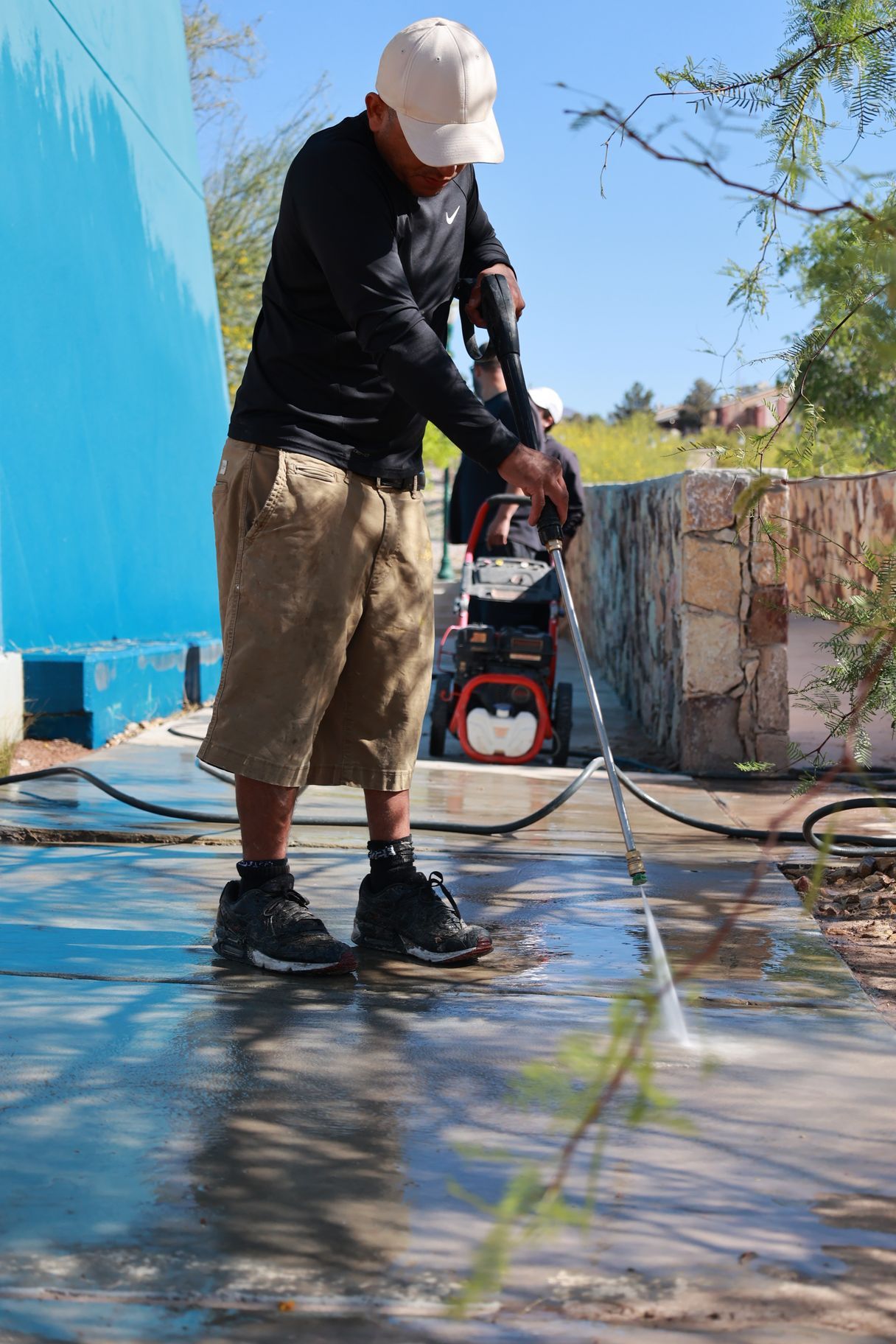 Worker pressure-washing a wet outdoor surface beside a blue wall under a bright sky