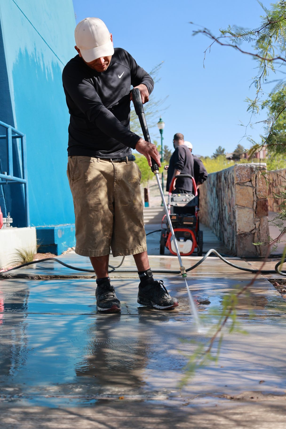 Person pressure-washes a wet concrete walkway beside a blue building on a sunny day.