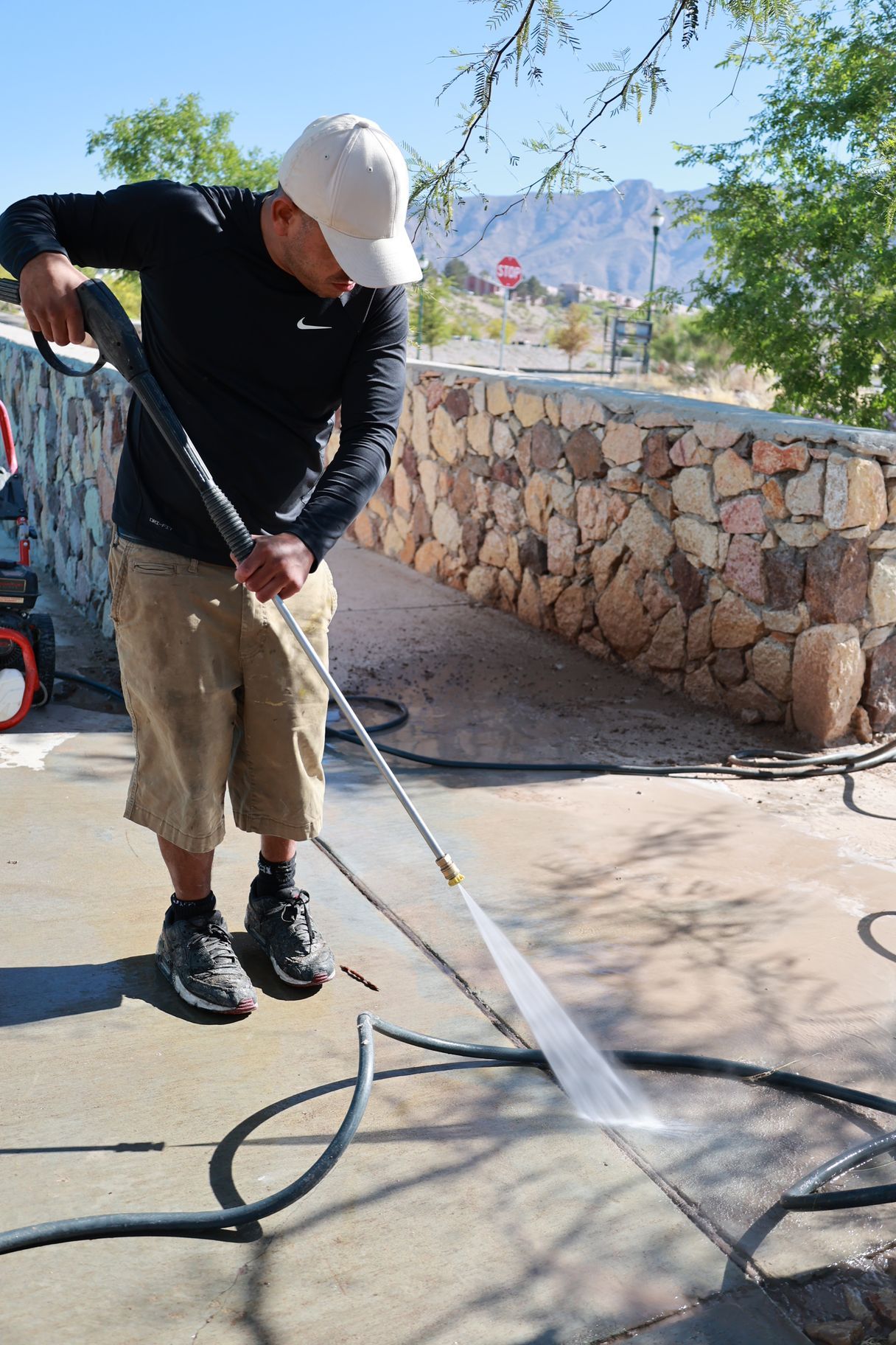 Person pressure-washing a concrete driveway beside a stone wall and tree on a sunny day