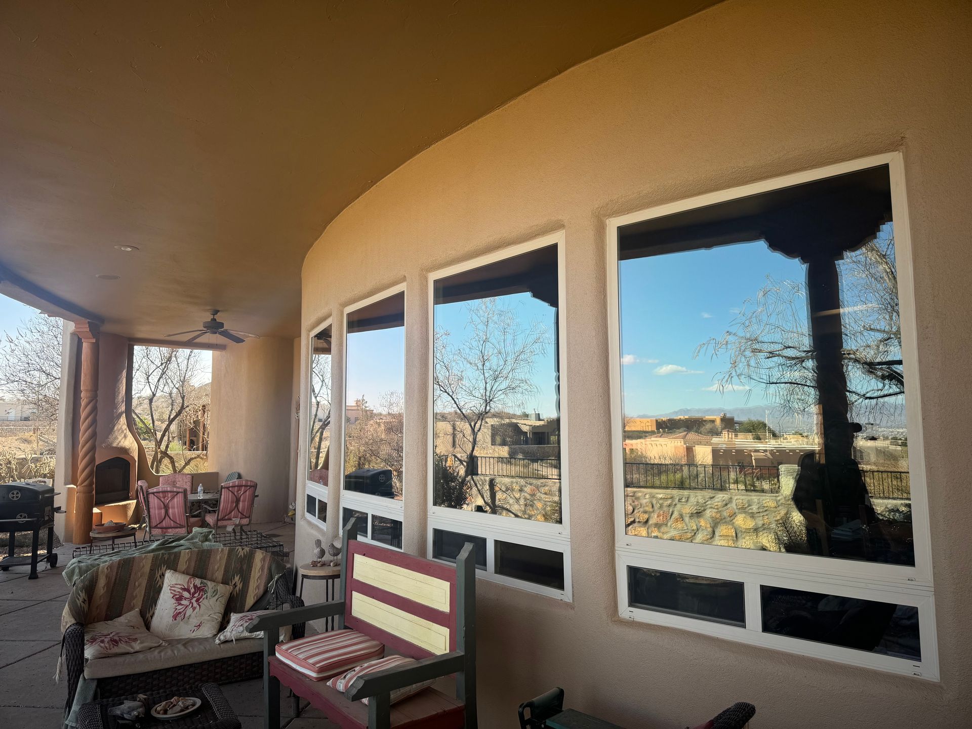 Covered patio with wicker chairs and a bench, overlooking a bright landscape through large windows.