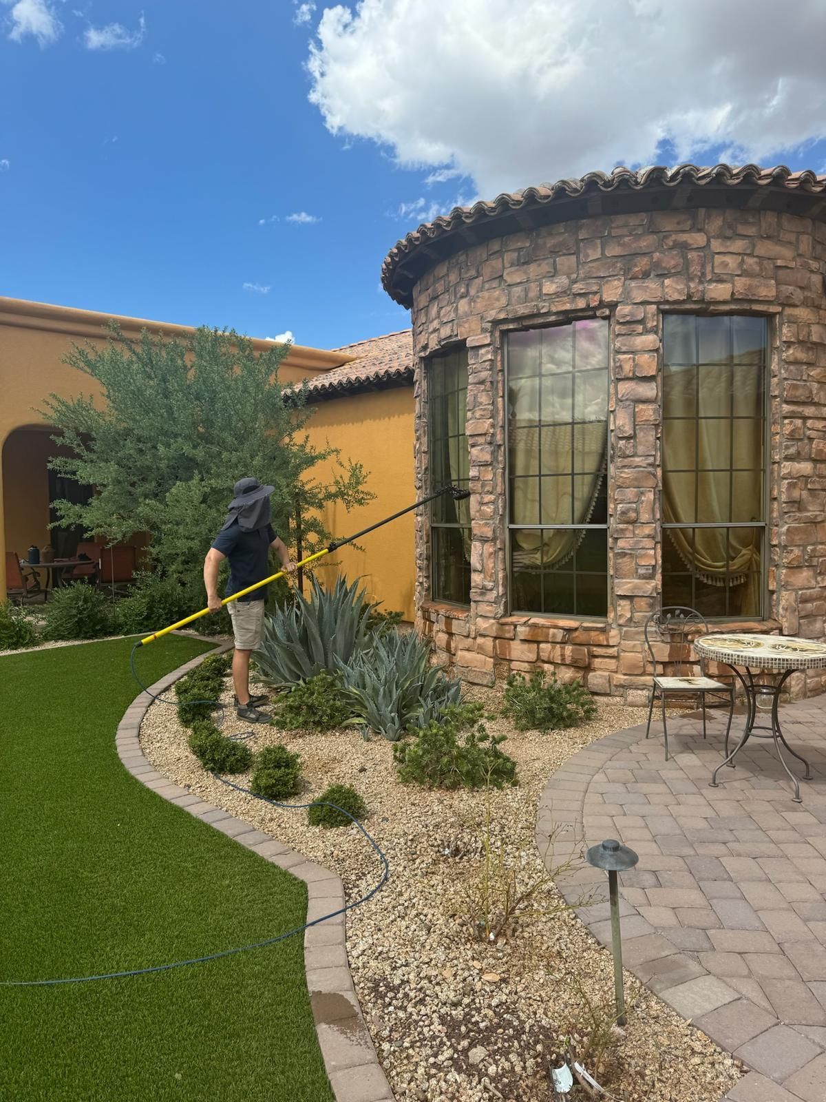 Person spraying a landscaped courtyard beside a stone building under a blue sky
