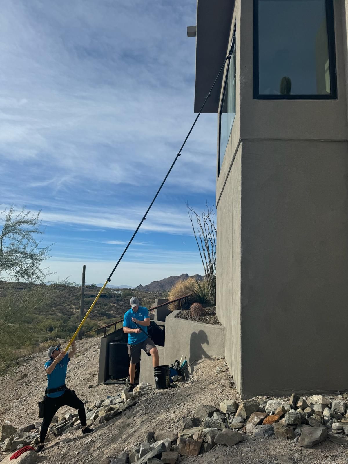Two people pulling a rope beside a house on a rocky slope under a blue sky.