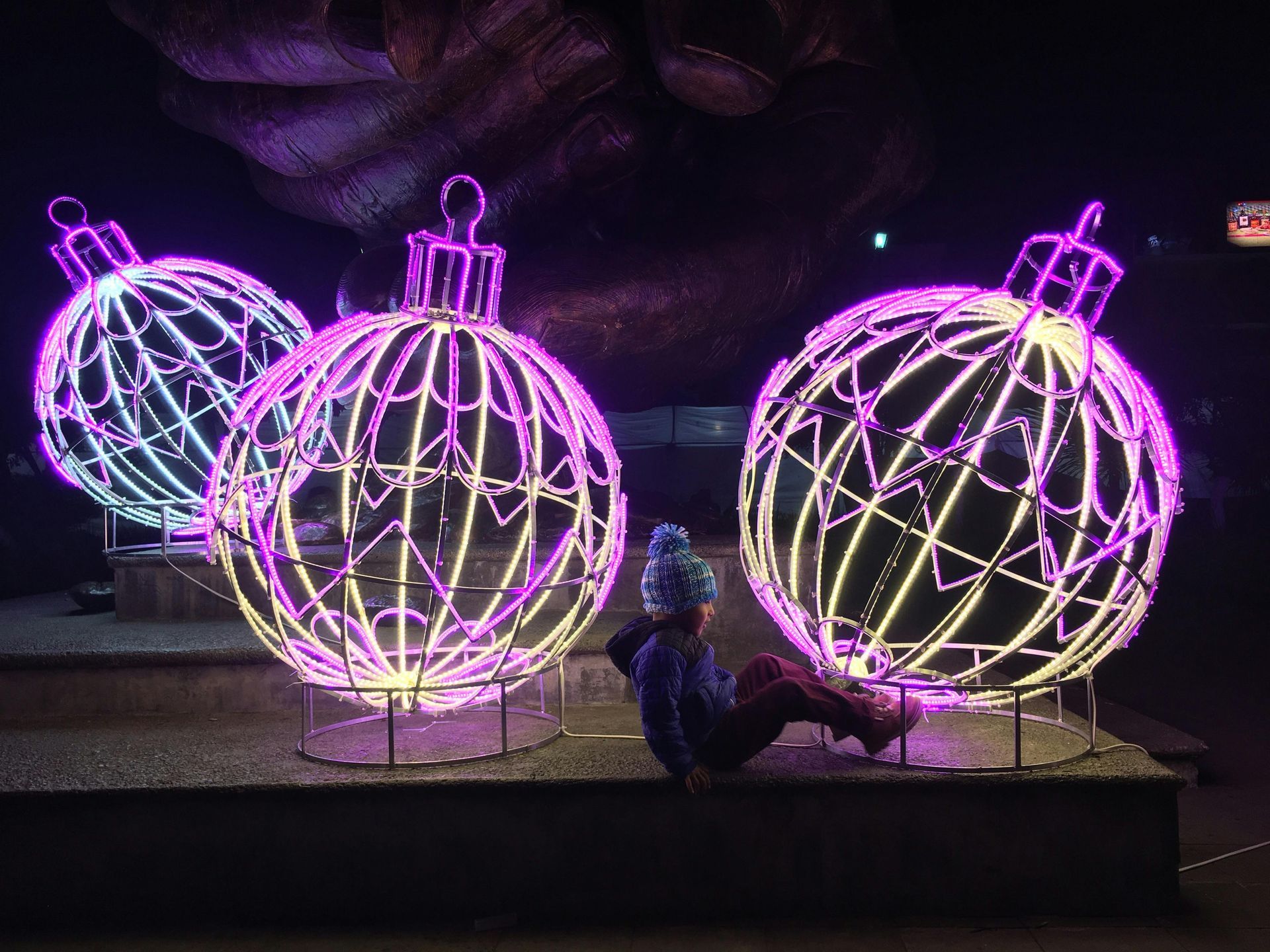 Purple and pink glowing holiday light sculptures shaped like ornaments on a nighttime display platform