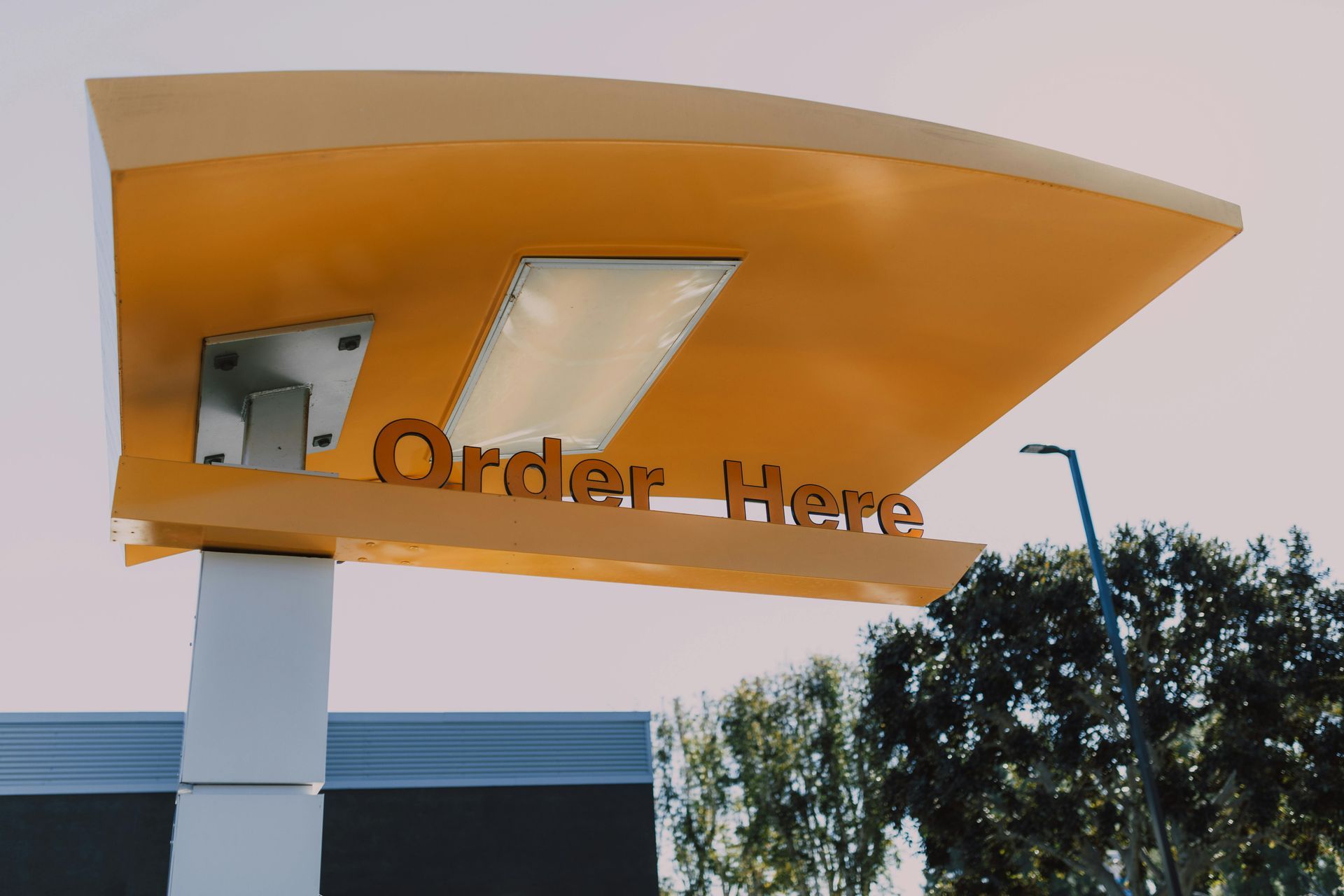 Orange drive-thru order canopy with an “Order Here” sign and a speaker box, against trees and sky