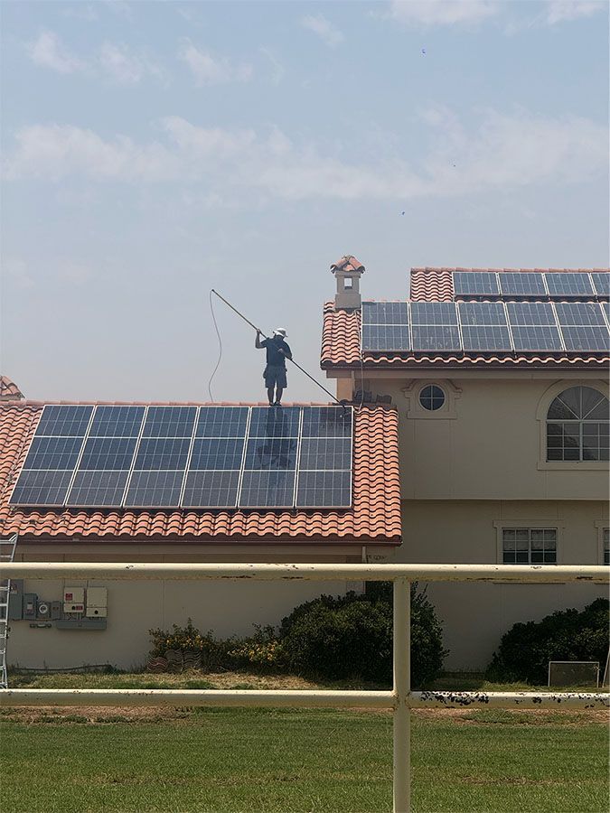 Worker cleaning rooftop solar panels on a house with red tile roof and blue sky.