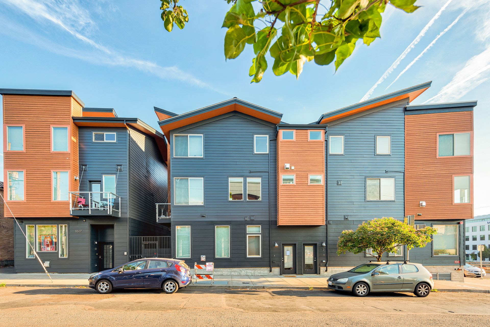 Modern townhouses with blue and brown siding, parked cars in front, sunny day.