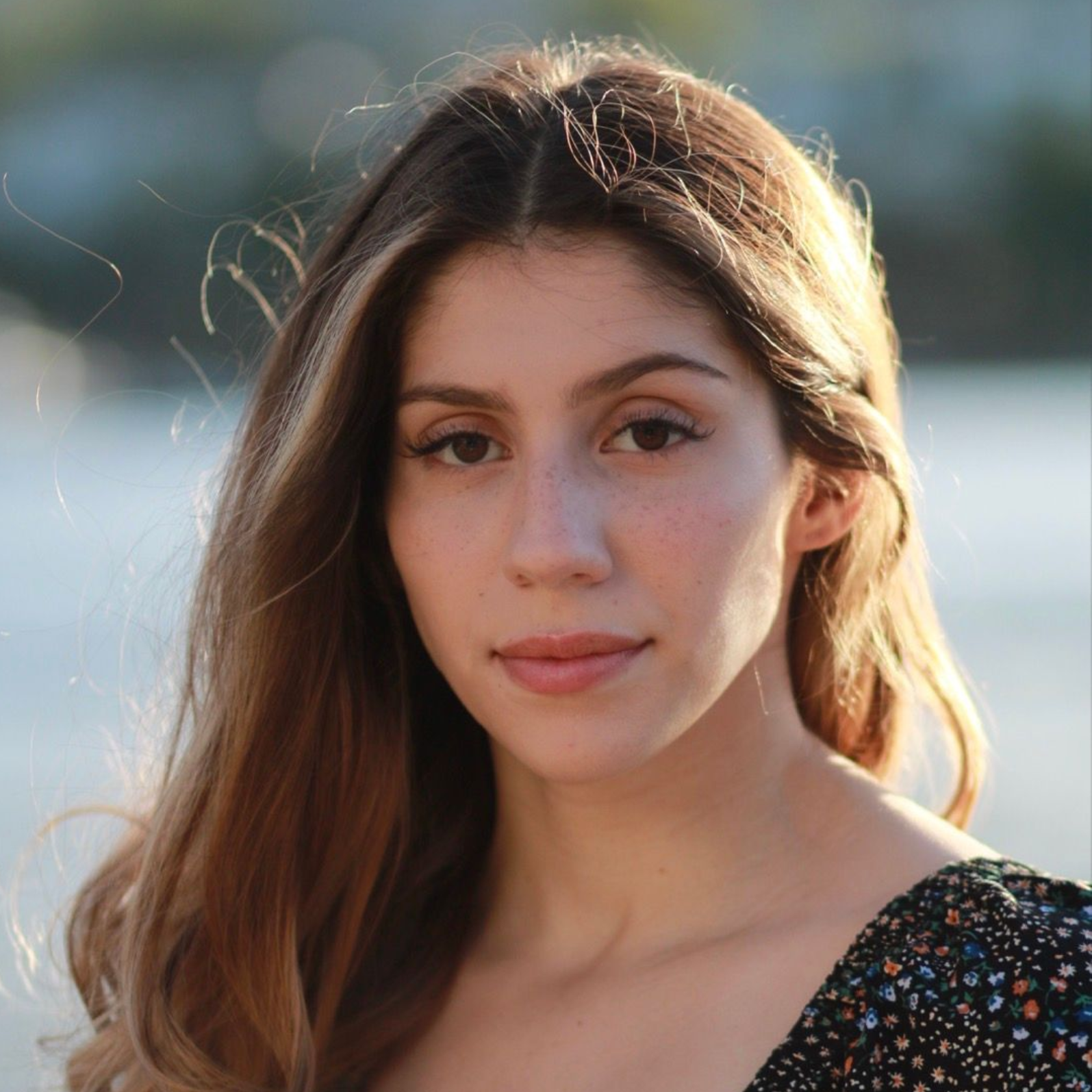 Portrait of a woman with long brown hair in a floral top, standing outdoors in soft sunlight.