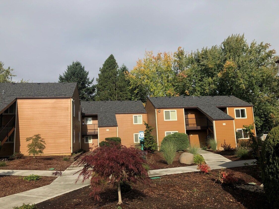 Apartment buildings with brown siding and dark gray roofs, surrounded by trees and landscaping.