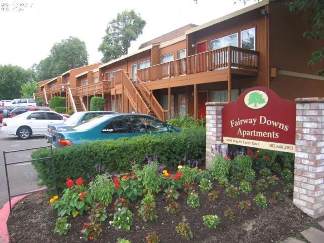 Fairway Downs Apartments, two-story brown building with balconies, green shrubs and flowers in front of sign.