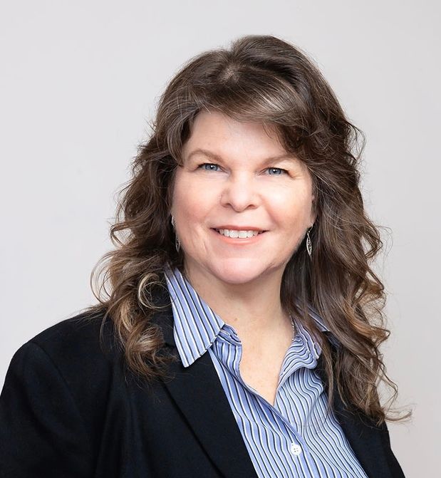 Woman with long, wavy brown hair, smiling, wearing a blue-striped shirt and black blazer.