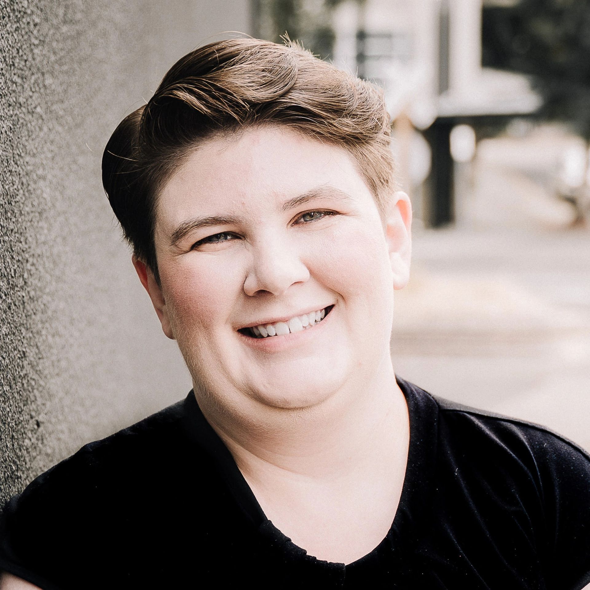 Smiling person in a black shirt standing beside a wall outdoors