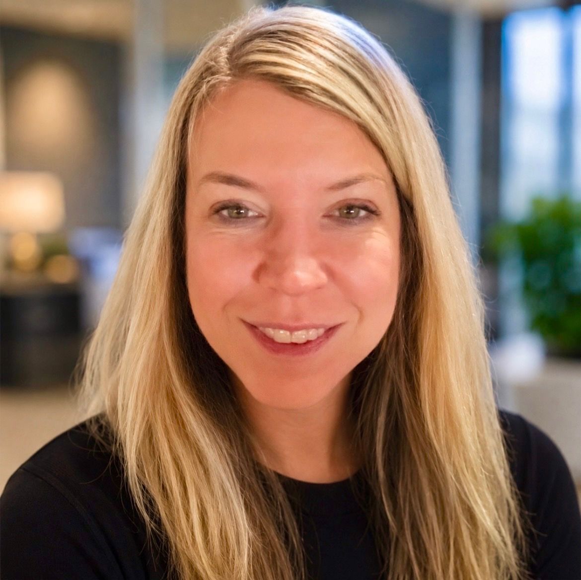 Smiling blonde woman in a black top, seated indoors with a softly blurred office background