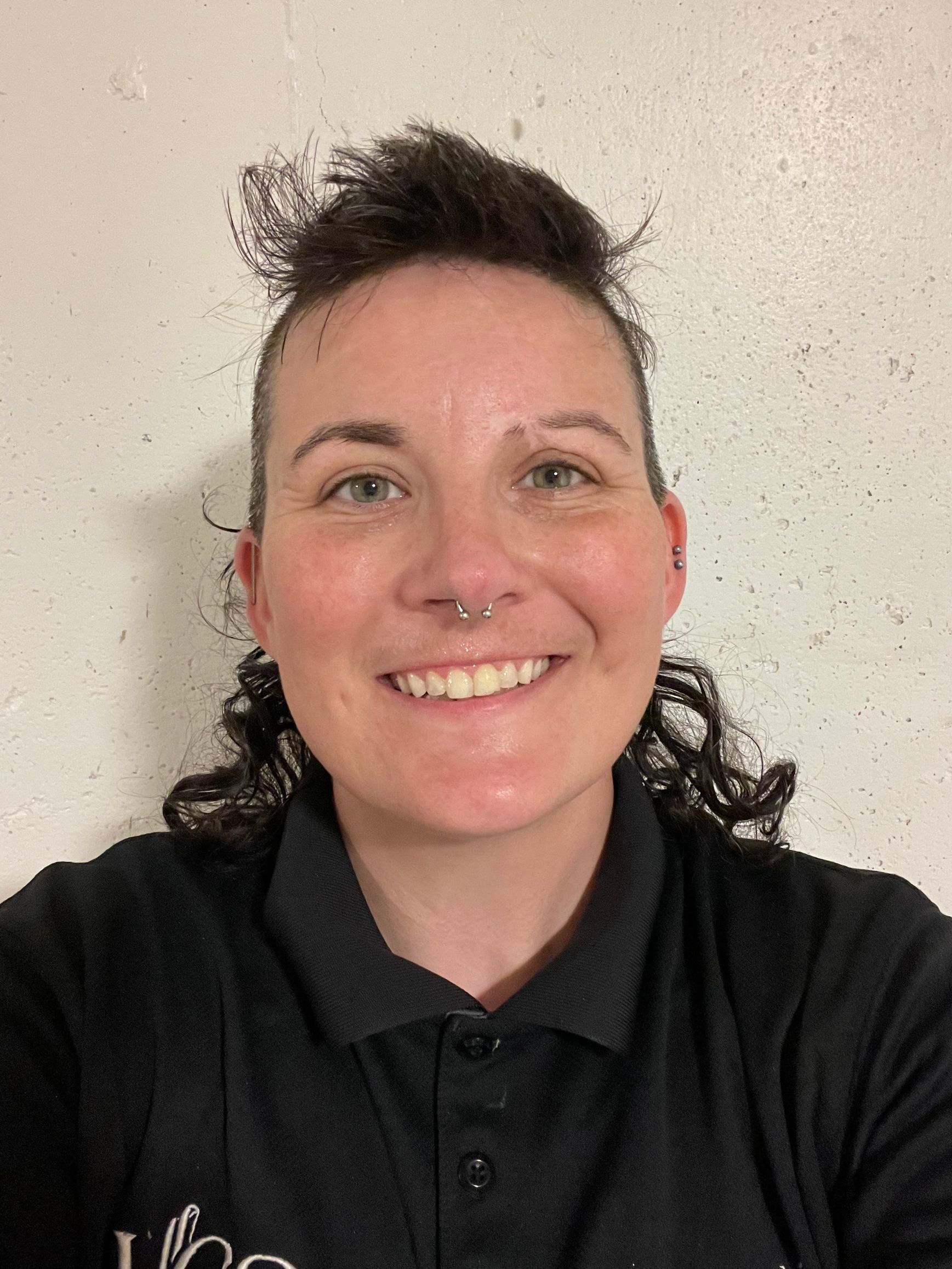 Smiling person in a black polo shirt against a light wall.