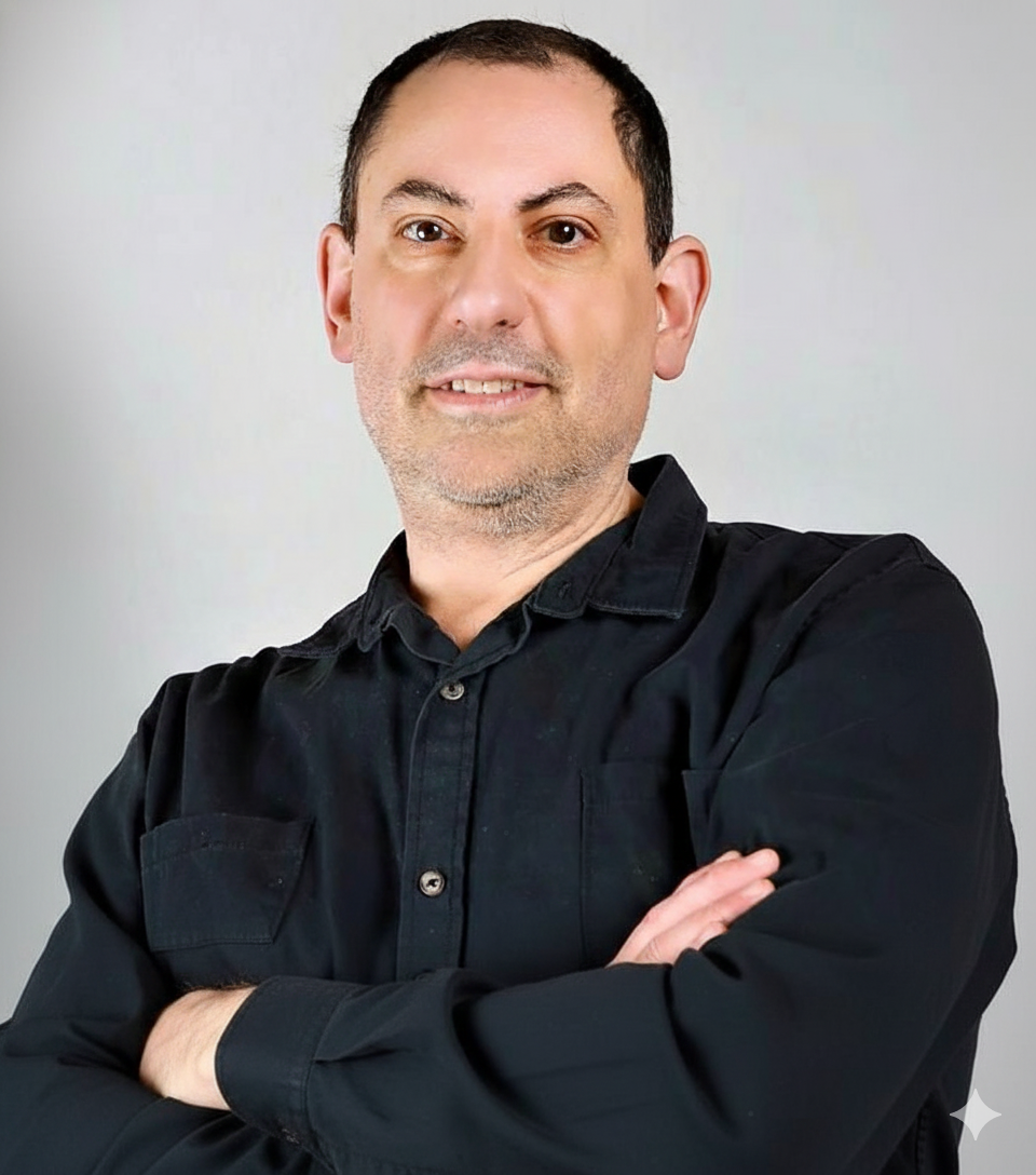 Man in a black button-up shirt with arms crossed, smiling, against a gray background.