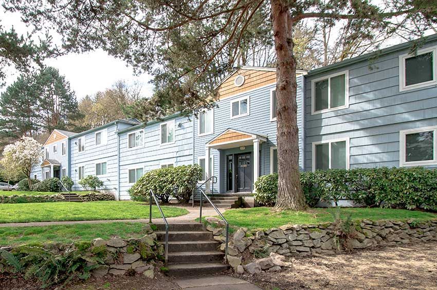 Apartment complex with light blue siding, green lawn, and stone retaining wall. A staircase leads to a central entrance.