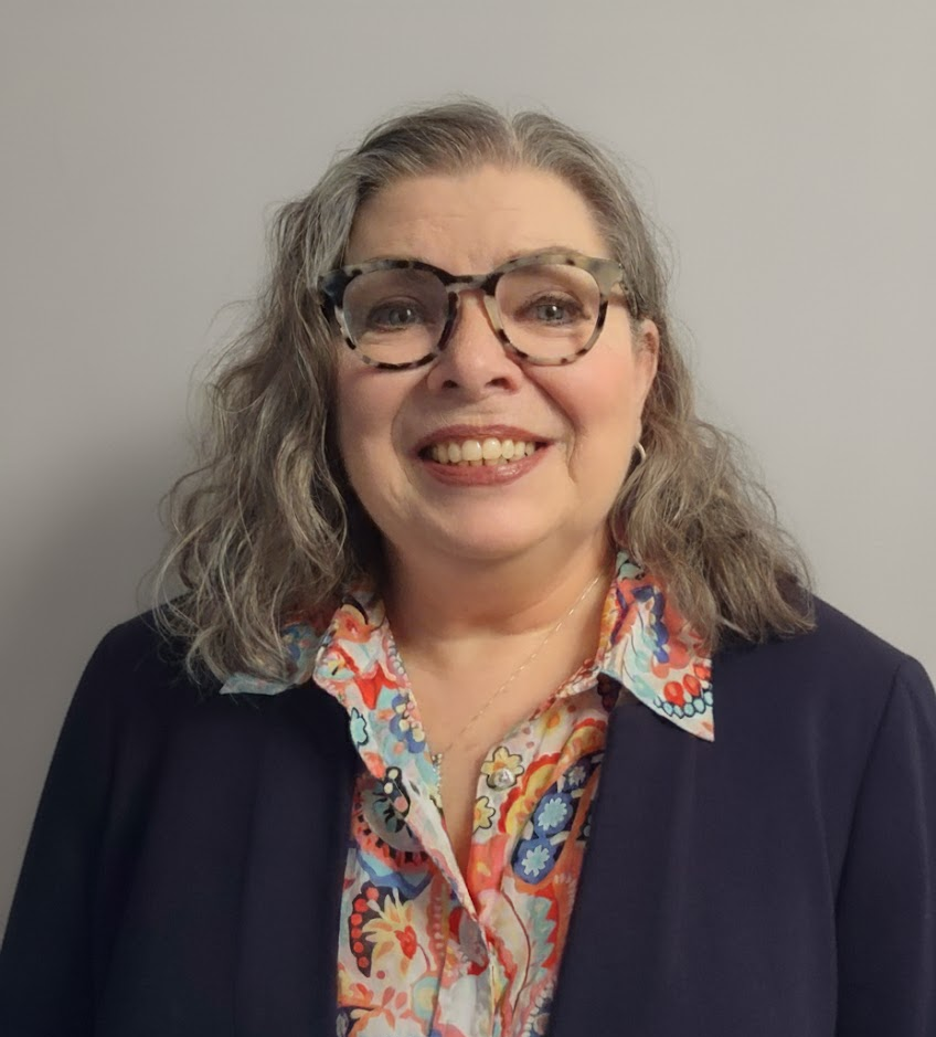 Woman with glasses smiling, wearing a patterned shirt and navy blazer against a gray backdrop.