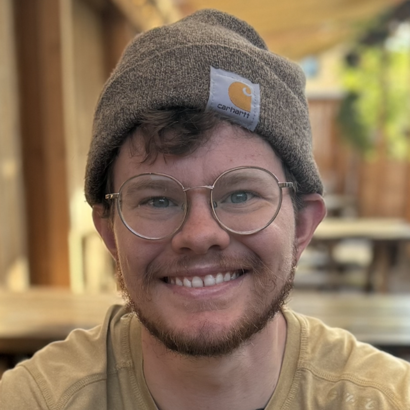 Smiling person wearing round glasses and a brown beanie in a warmly lit indoor setting