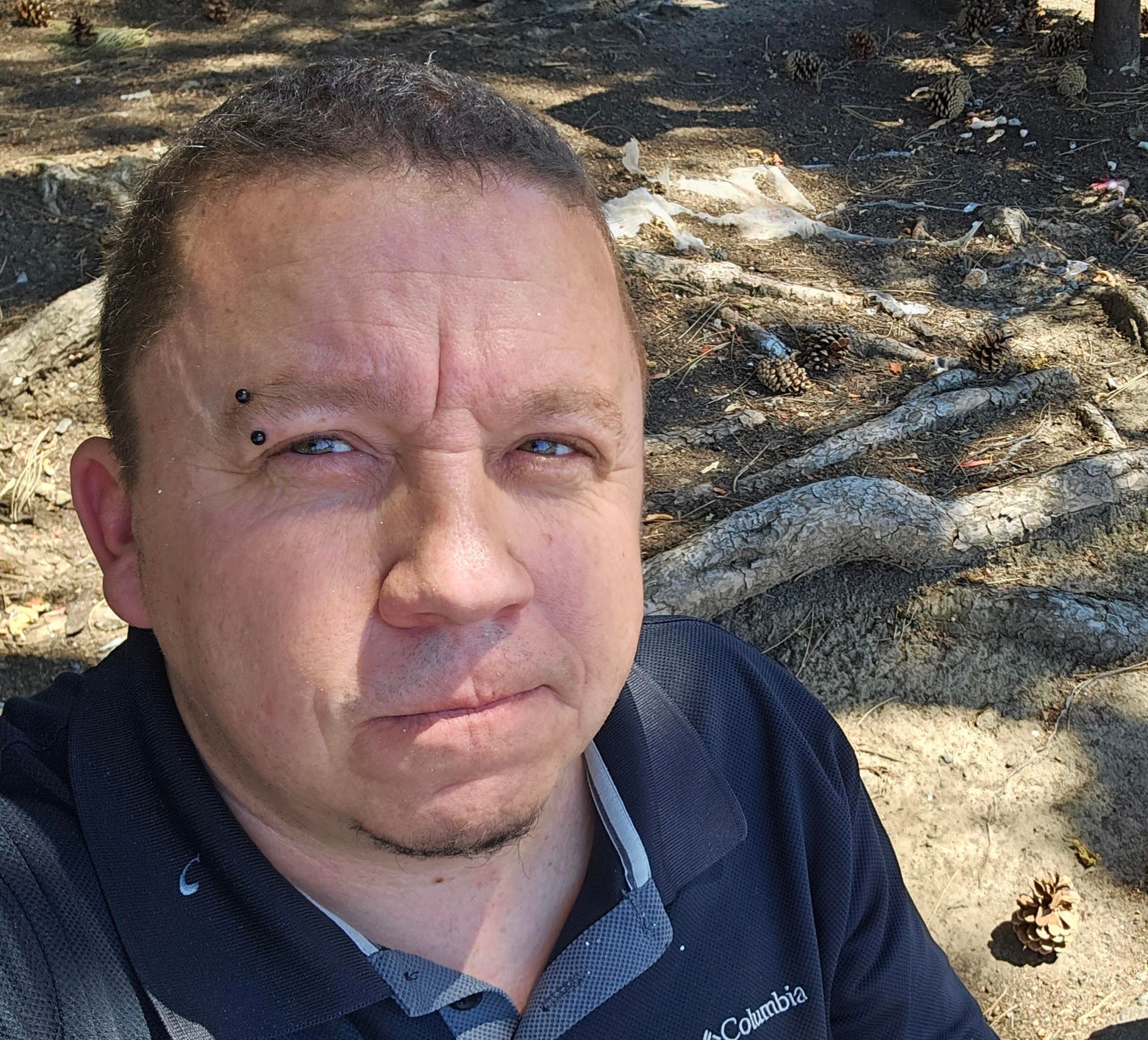 Close-up selfie of a man outdoors in dappled sunlight, wearing a dark polo shirt, with a neutral expression