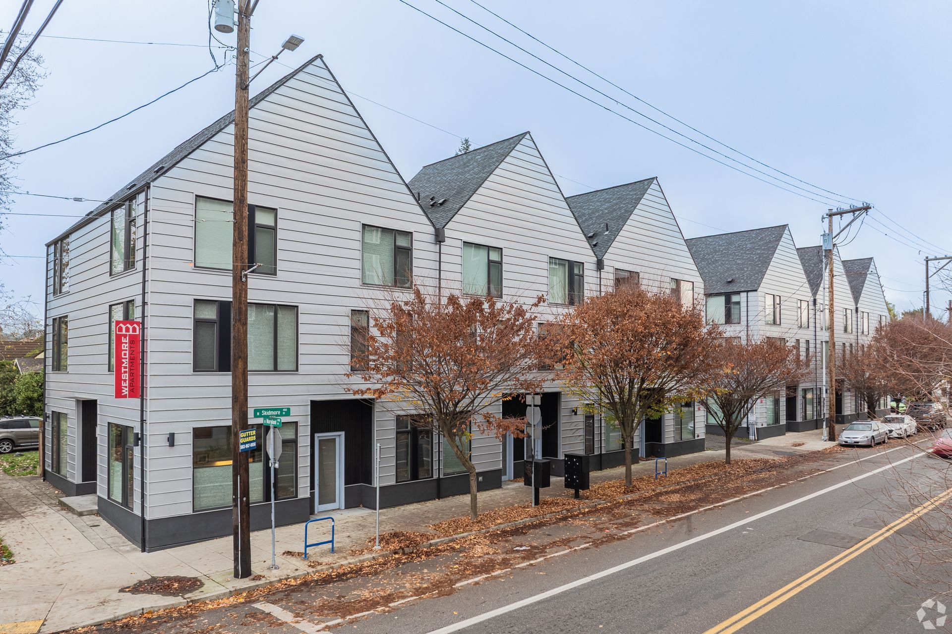 Row of modern, light gray townhouses with black accents and gabled roofs along a street with bare trees.